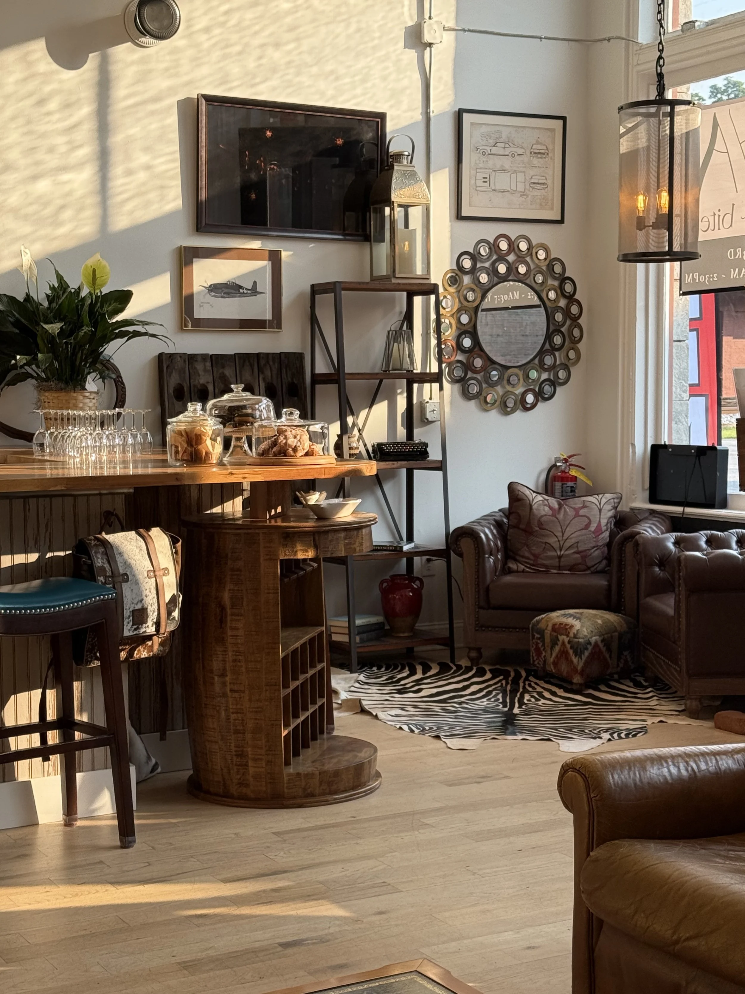 Interior of a cozy living room with a wooden bar, leather armchairs, a zebra-patterned rug, decorative wall art, a potted plant, and a window letting in sunlight.