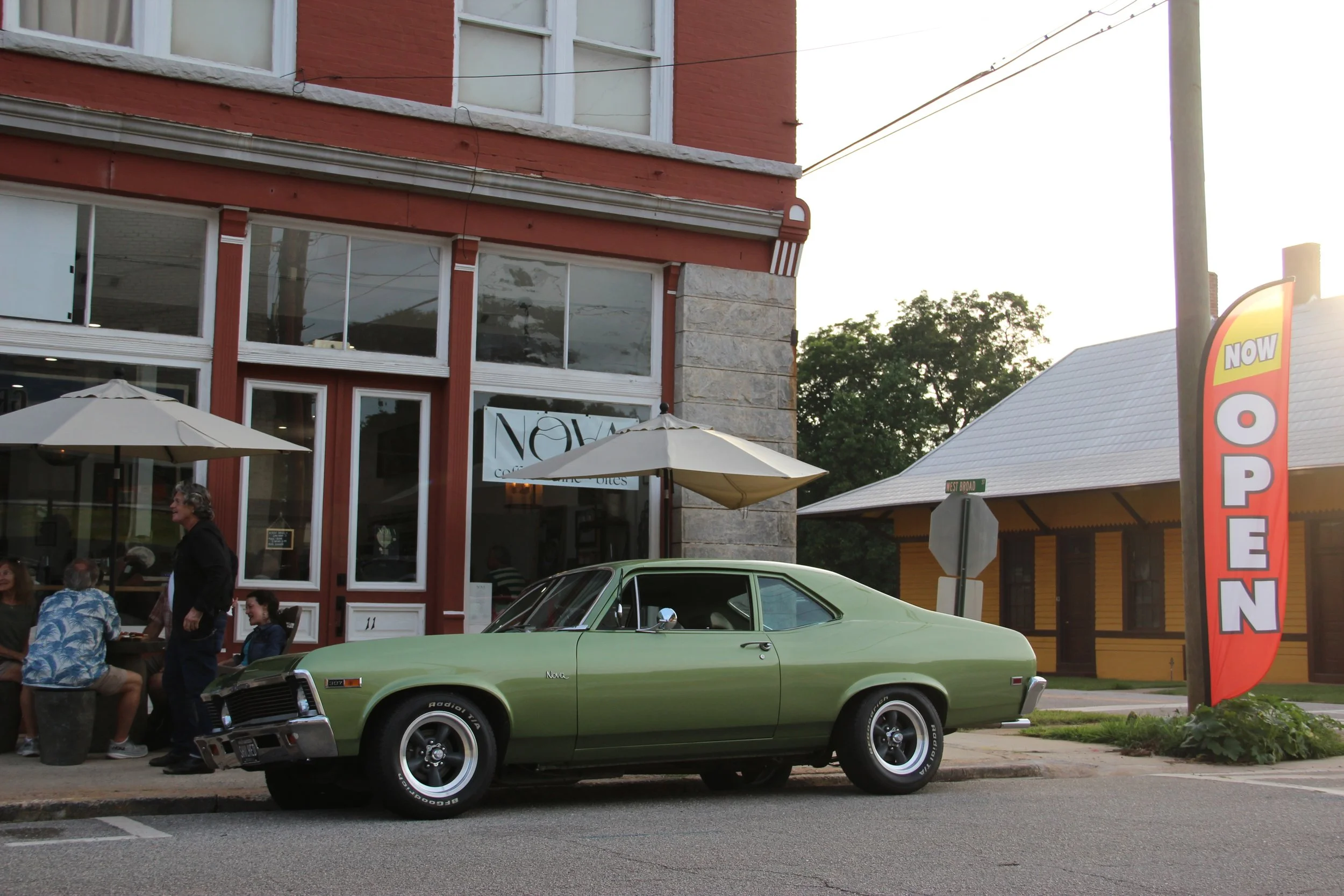 A vintage green car parked in front of a restaurant with outdoor seating and a large red "Open" sign on a signpost. People are dining outside under umbrellas.