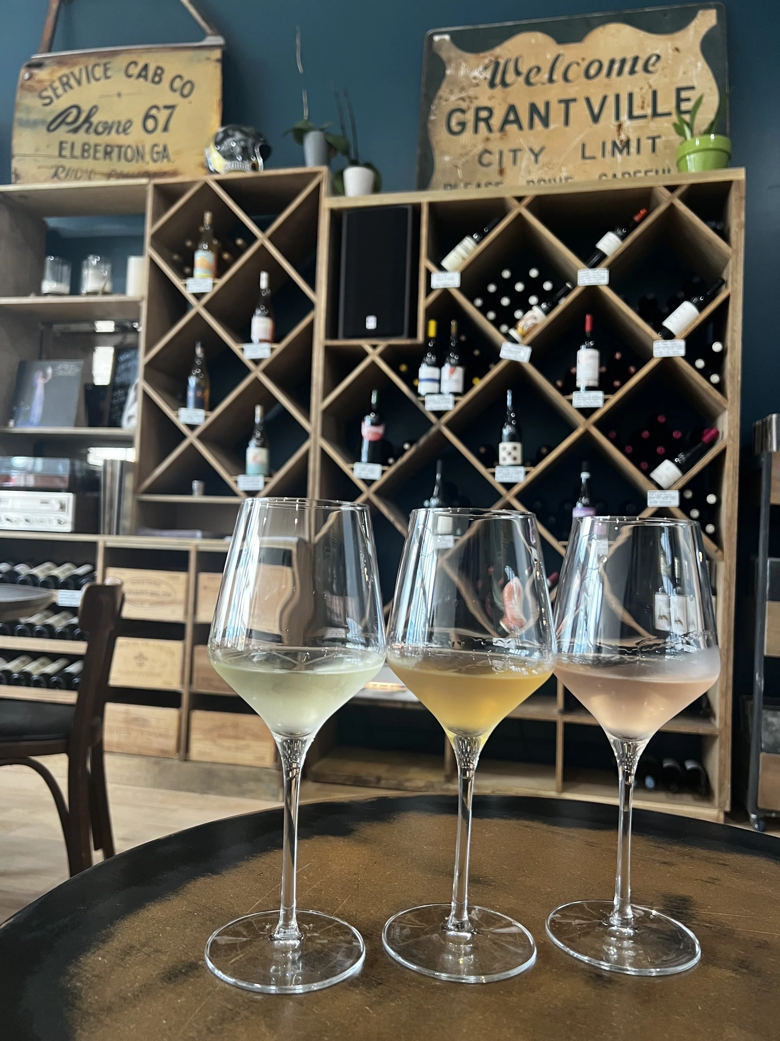 Three wine glasses filled with white, yellow, and pink wine on a wooden table inside a wine shop or tasting room.
