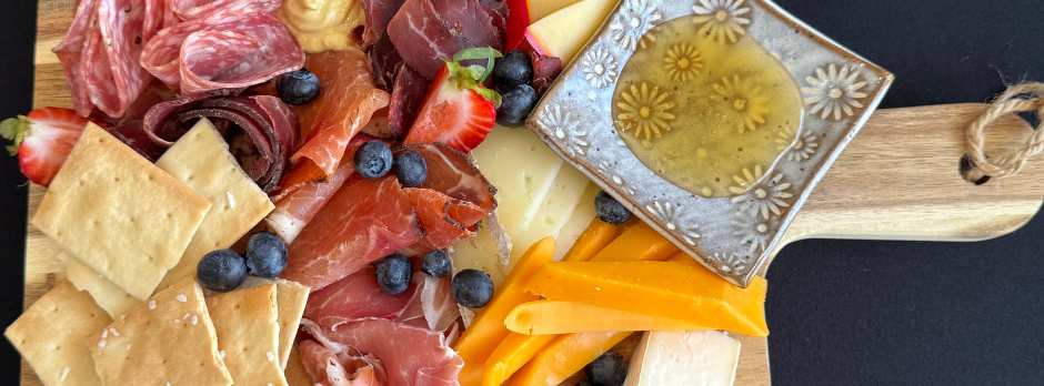 Cheese, crackers, strawberries, blueberries, charcuterie meats, honey in a decorative dish, and orange slices on a wooden serving board.