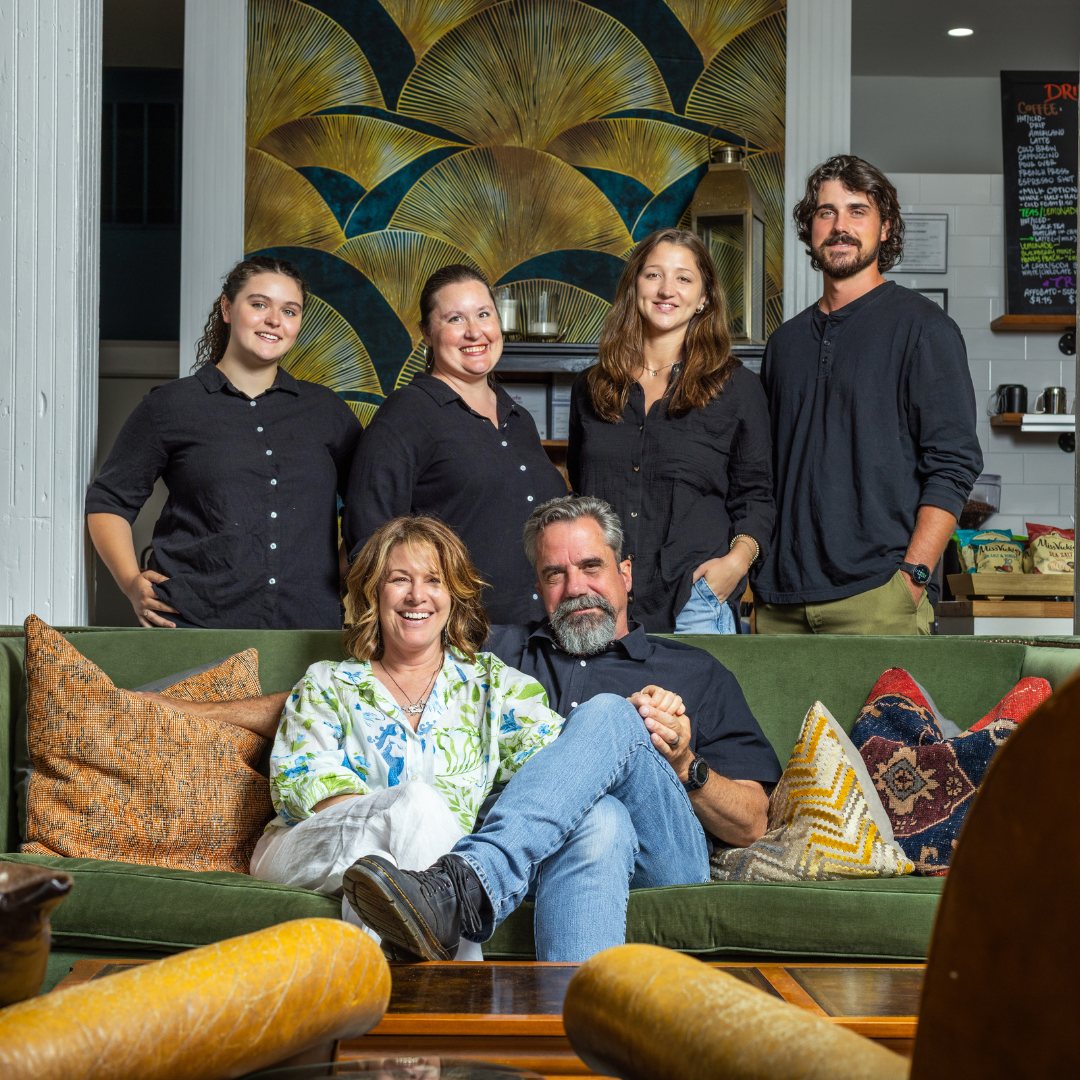 A group of six people, three women and three men, posing together in a cozy indoor setting with colorful pillows on a green sofa and a decorative gold and blue patterned wall background.