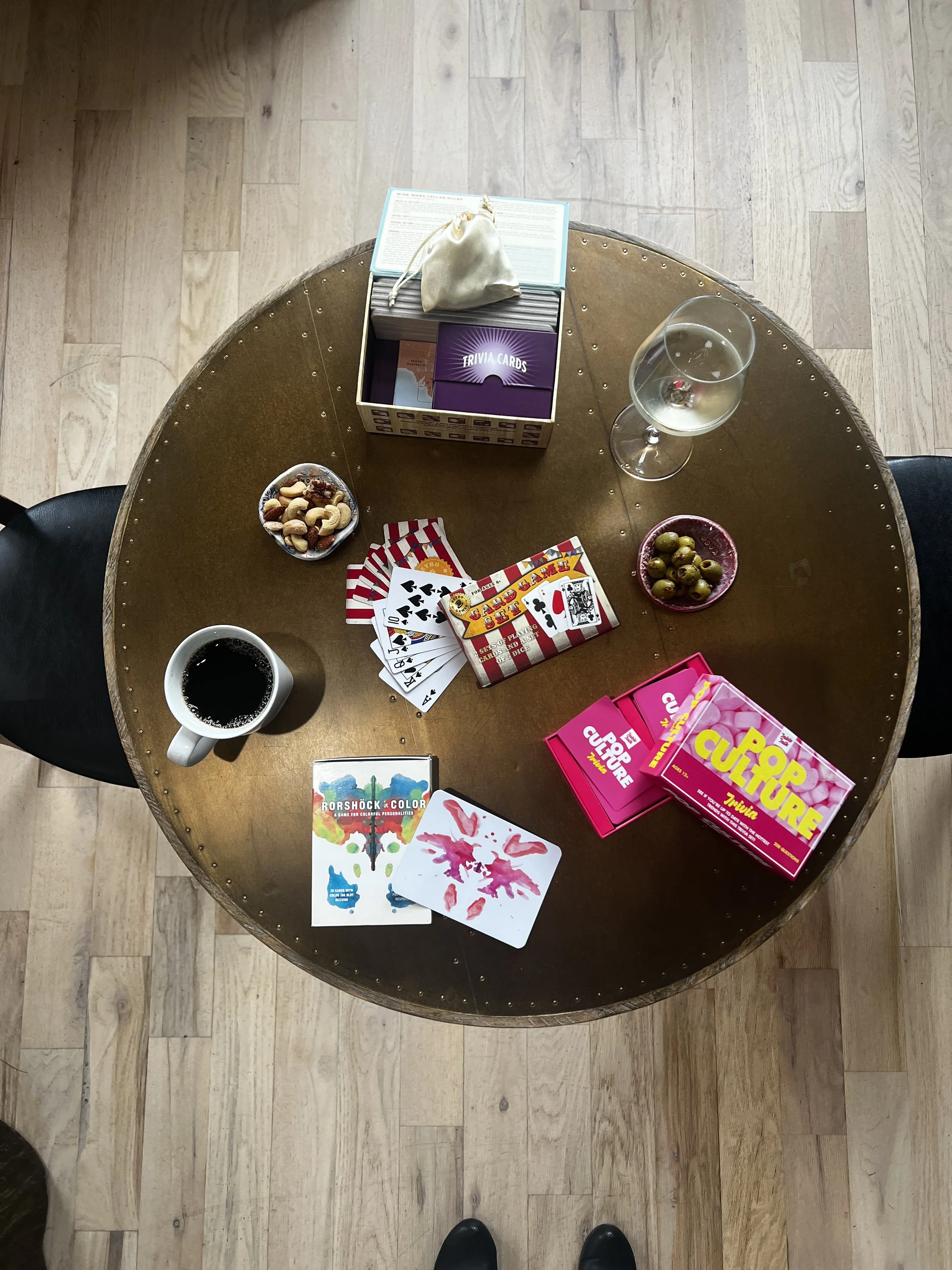 Top-down view of a round wooden table with various items including a white mug of black coffee, a glass of white wine, a bowl of mixed nuts, a bowl of green olives, a box of trivia cards, two boxes of card games, and some game cards, with black chairs and light wooden flooring surrounding the table.