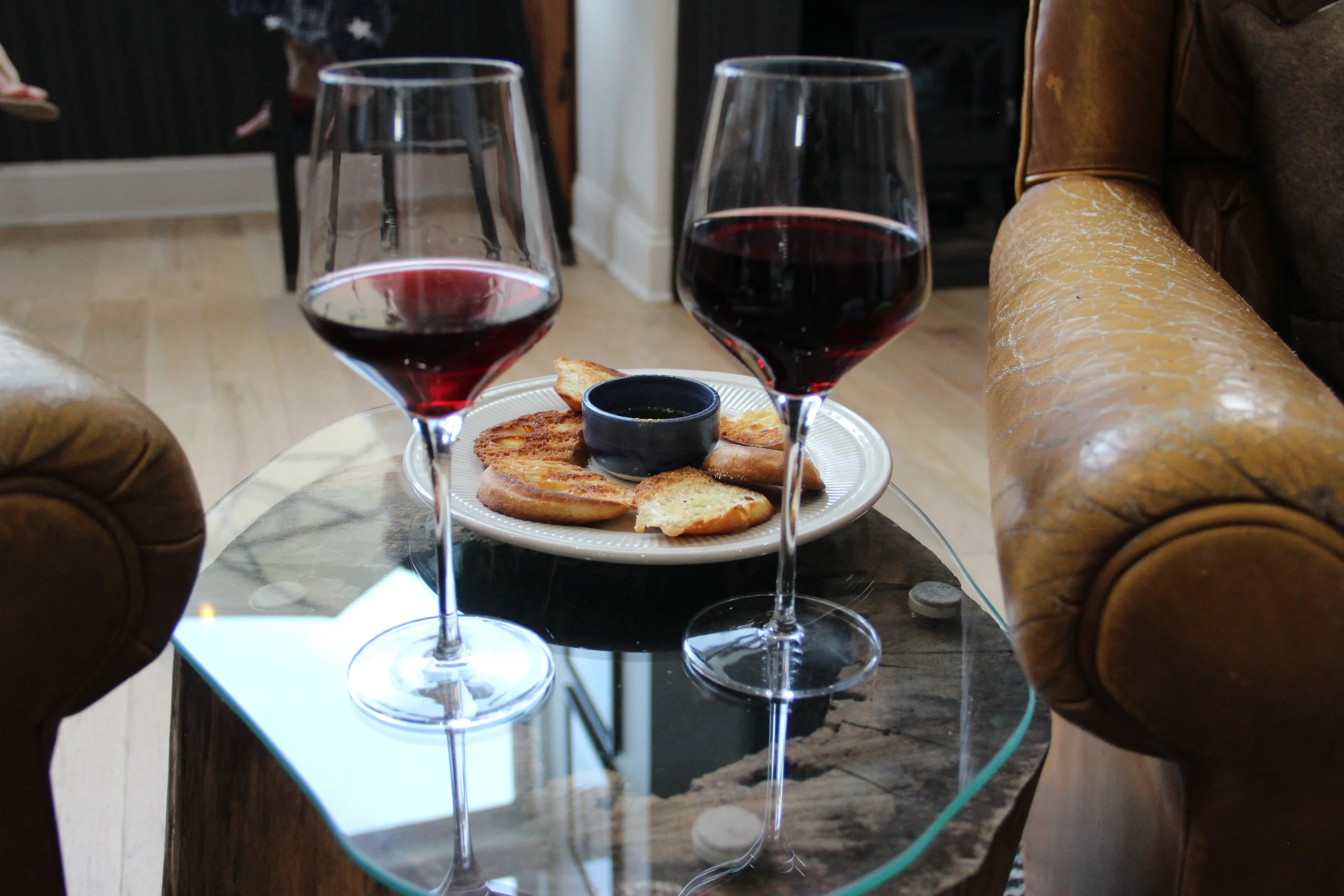 Two glasses of red wine on a glass table, with a plate of toasted bread slices and a small bowl of dipping sauce in between.