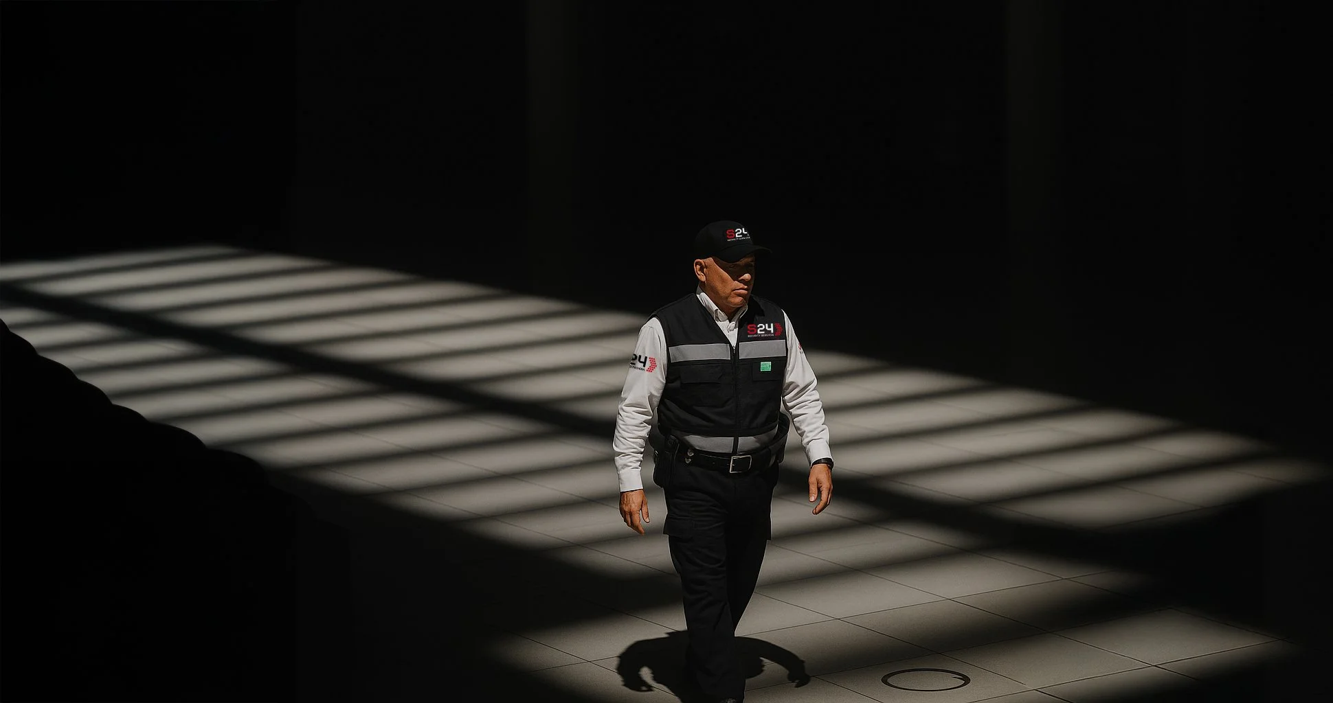 Hombre con uniforme y gorra caminando en un espacio oscuro con sombras de líneas en el suelo.
