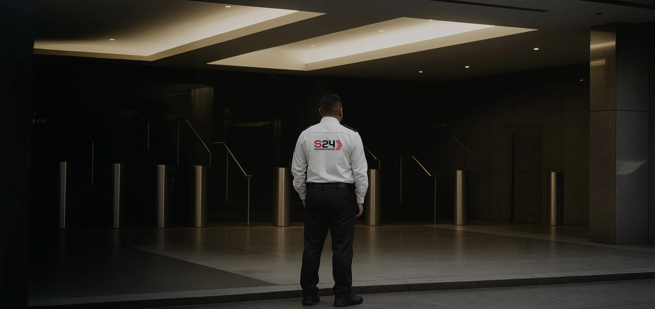 Vigilante con uniforme de S24 Security Seguridad Privada observa un vestíbulo oscuro con barandillas doradas.