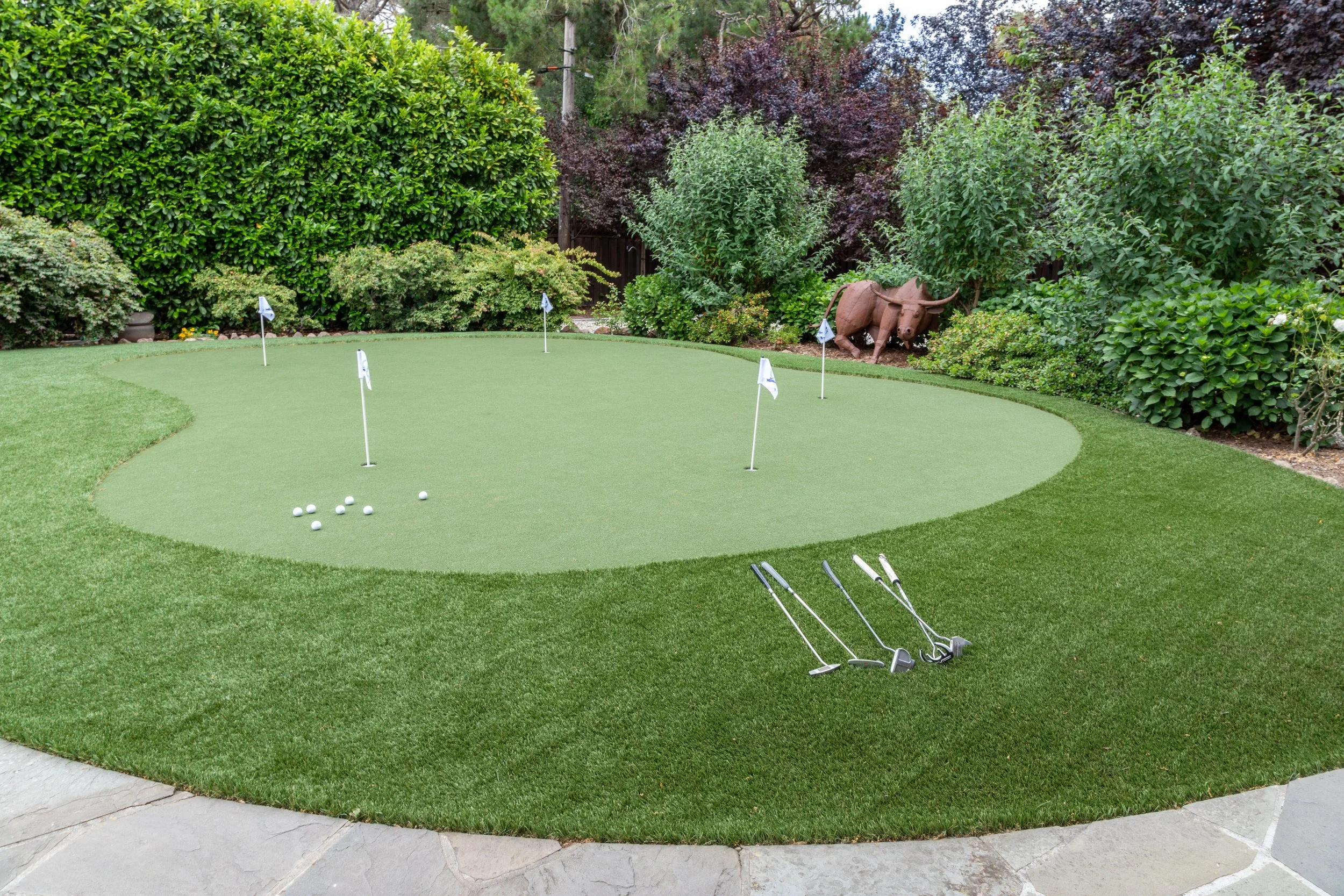A backyard putting green with five golf flags, a few golf balls, and golf clubs resting on the grass. There are lush bushes and trees in the background, and a rusted, decorative sculpture of a buffalo among the plants.