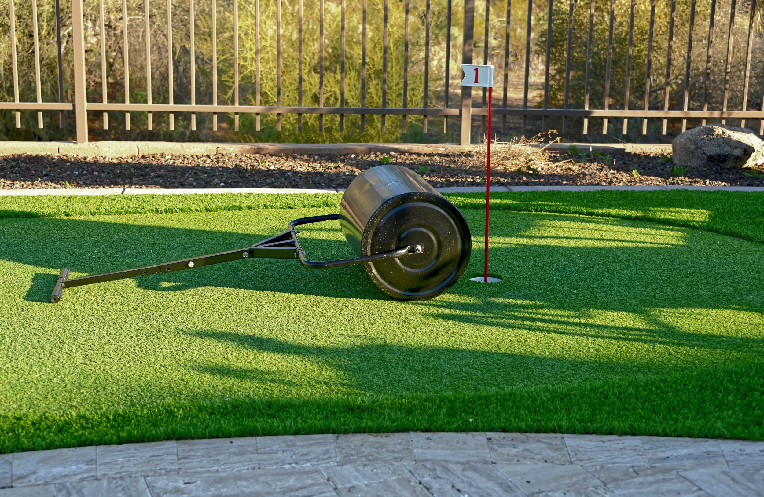 Miniature golf putting green with metal rolling cart, next to a flag marked with the number 1, surrounded by a stone border and a wooden fence in the background.