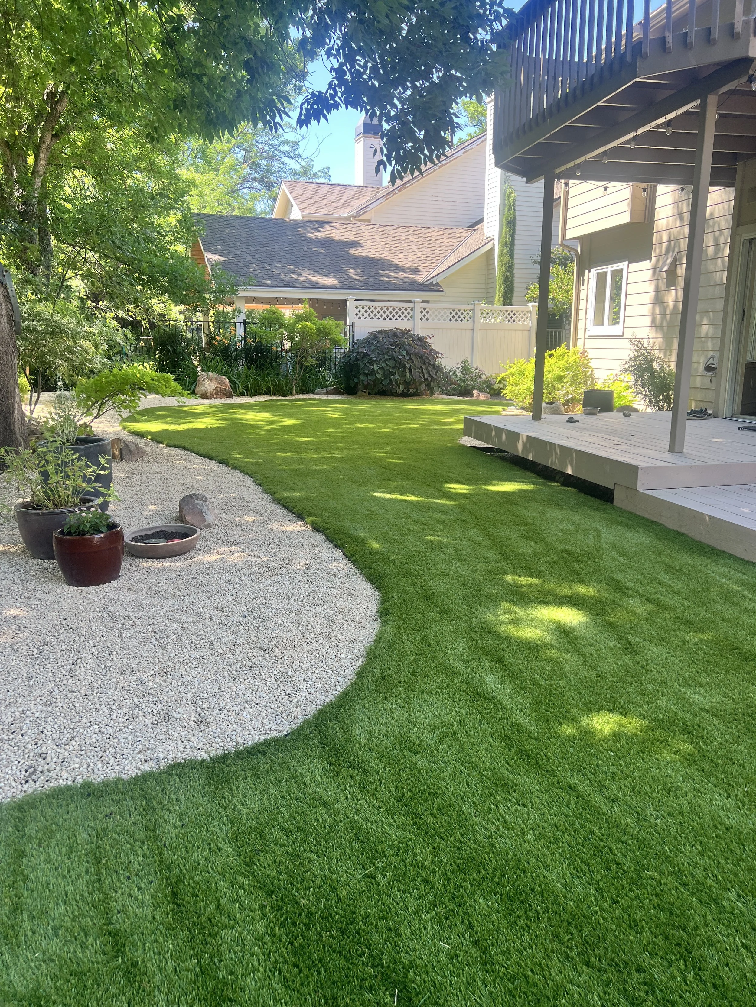 A backyard with a well-maintained green lawn, some potted plants on a gravel pathway, a deck attached to a house, and trees providing shade.
