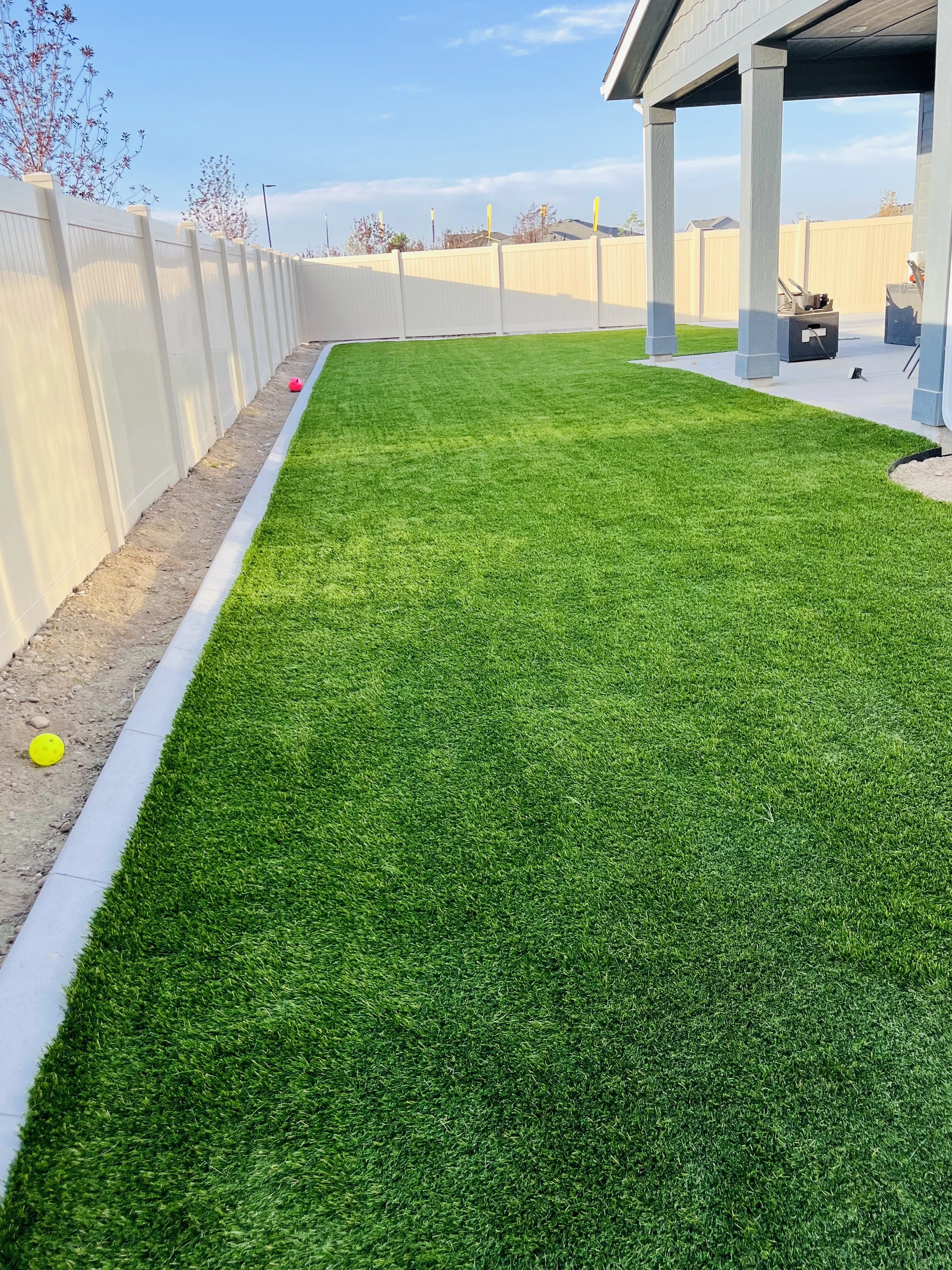 A backyard with a well-maintained green lawn, a white vinyl fence on the left, a covered patio with outdoor furniture on the right, and a blue sky with some clouds.