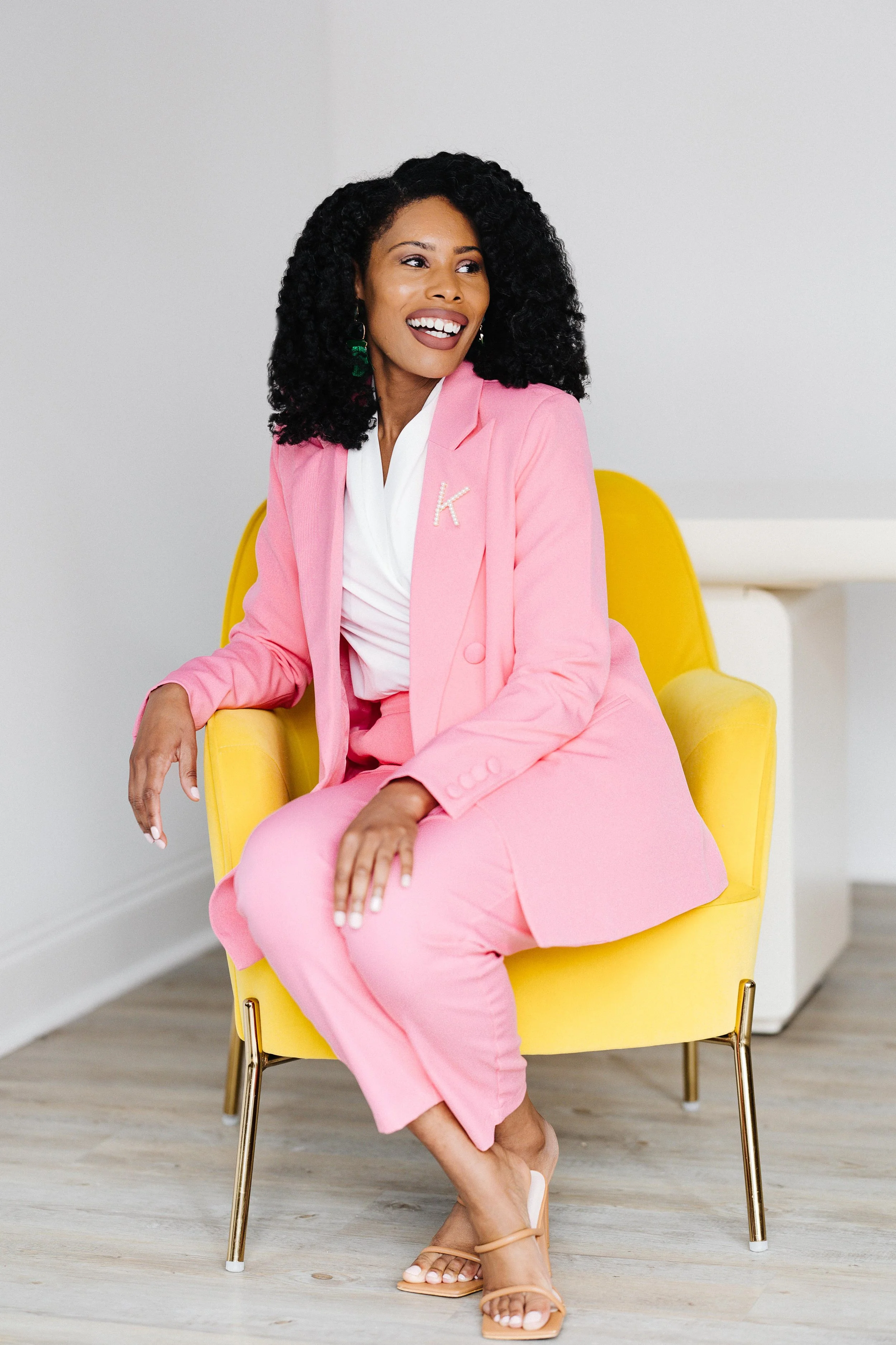 A woman with curly black hair, wearing a pink suit and white top, is sitting on a yellow armchair. She is smiling and looking to the side.