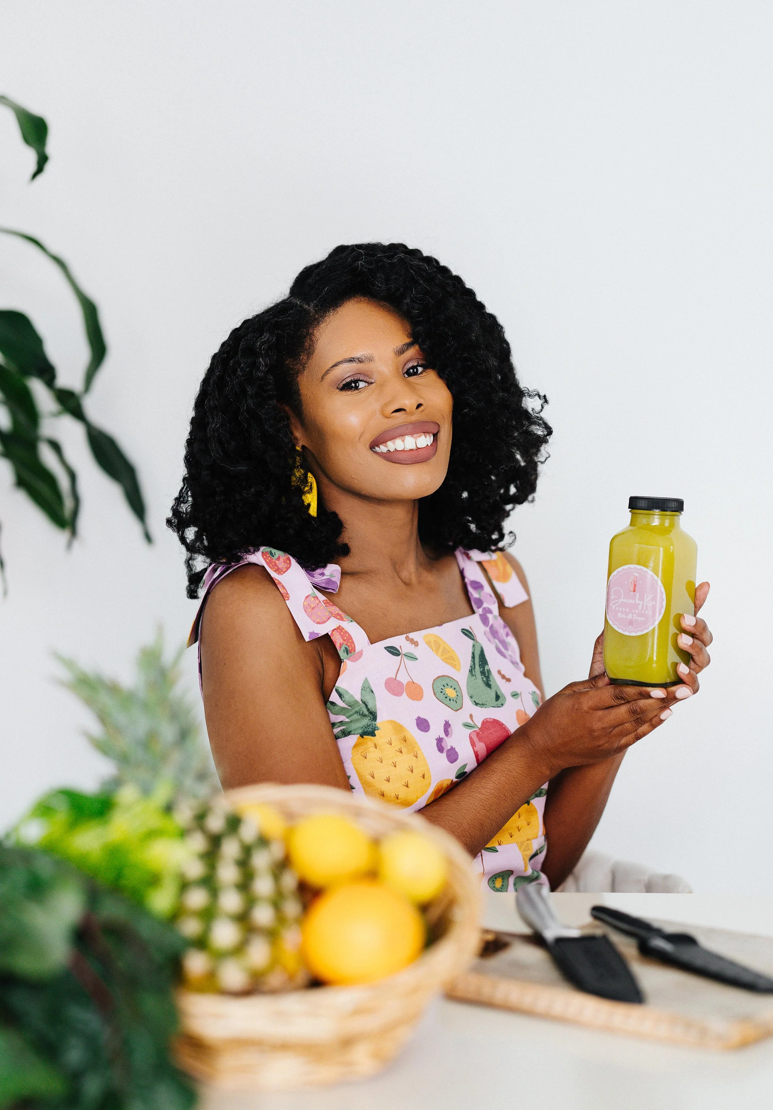 A woman with curly black hair, wearing a colorful fruit-patterned dress, is smiling and holding a yellow juice bottle with a pink sticker. There is a fruit basket with pineapples, lemons, and grapes in the foreground, and the background is plain white.