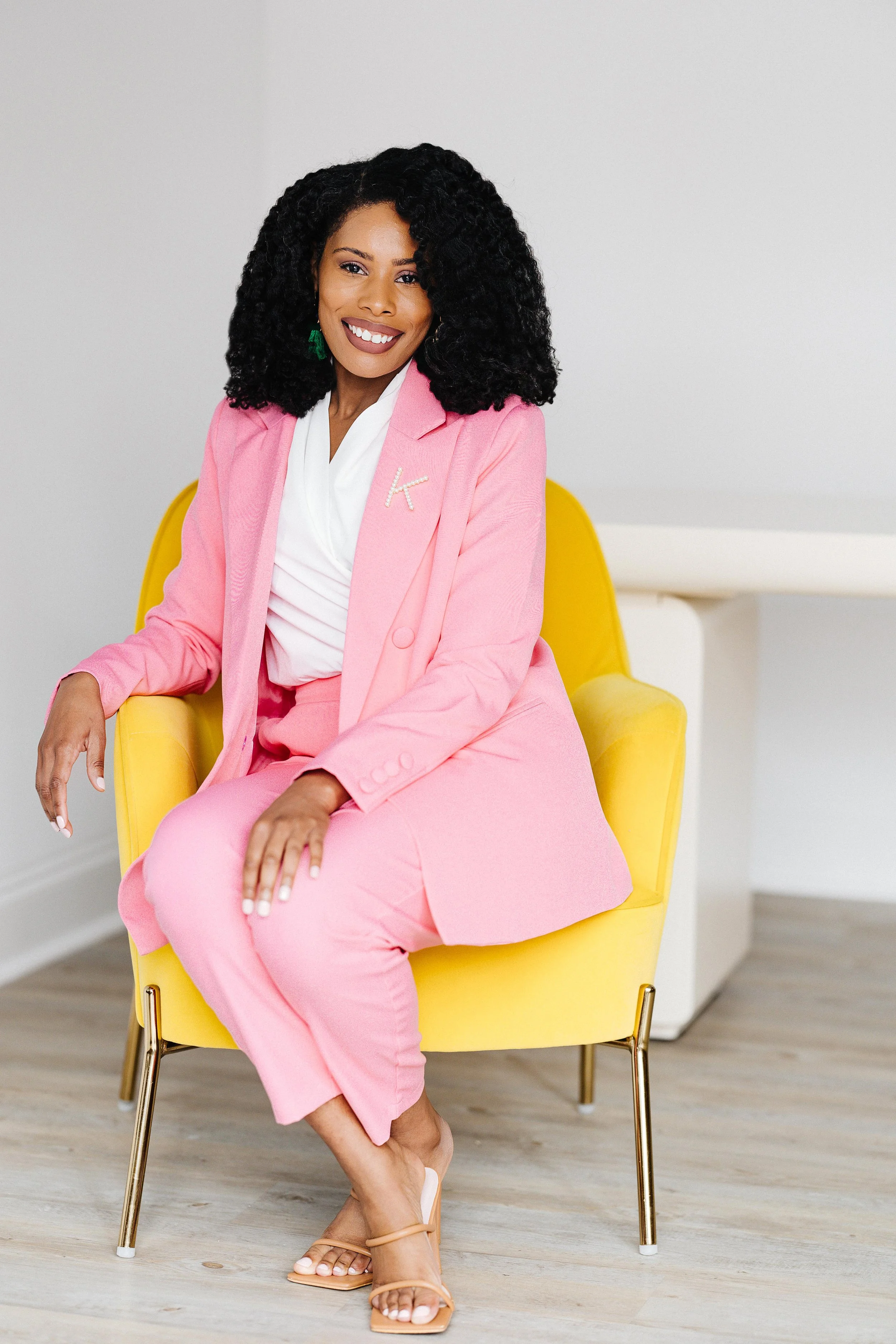 A woman with curly black hair, wearing a pink suit with a white top, sitting on a yellow chair, smiling with her legs crossed, in a minimalistic room.