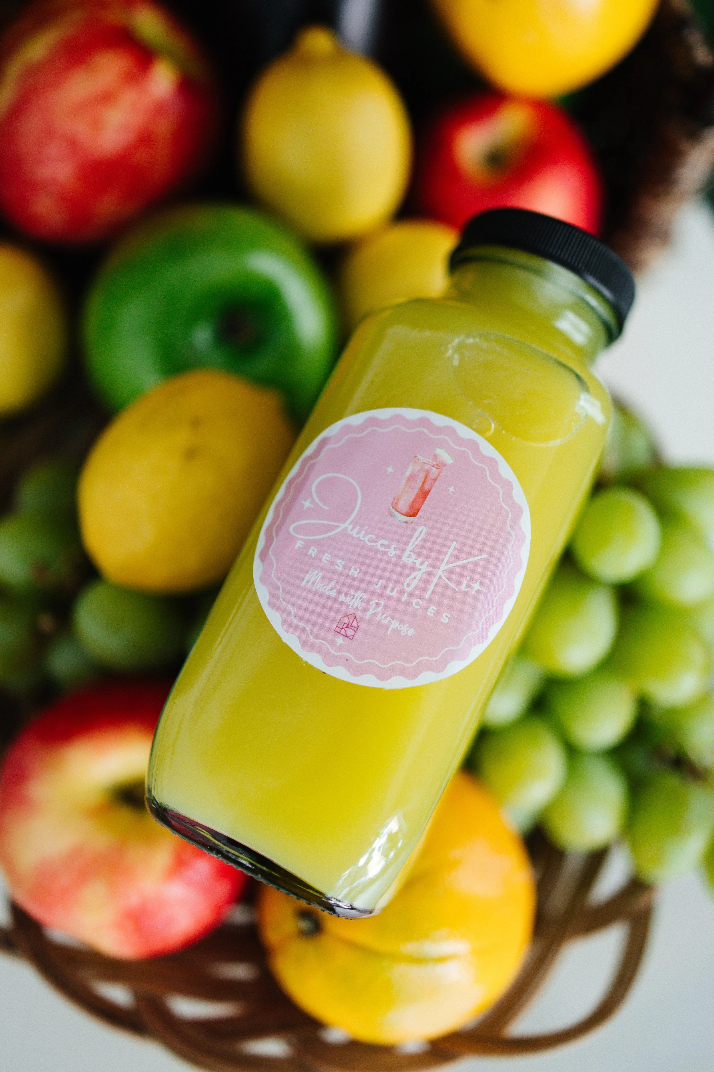 A bottle of yellow juice labeled "Juices by Kx Fresh Juices Made with Purpose" is placed in a basket filled with green and red apples, lemons, and grapes.