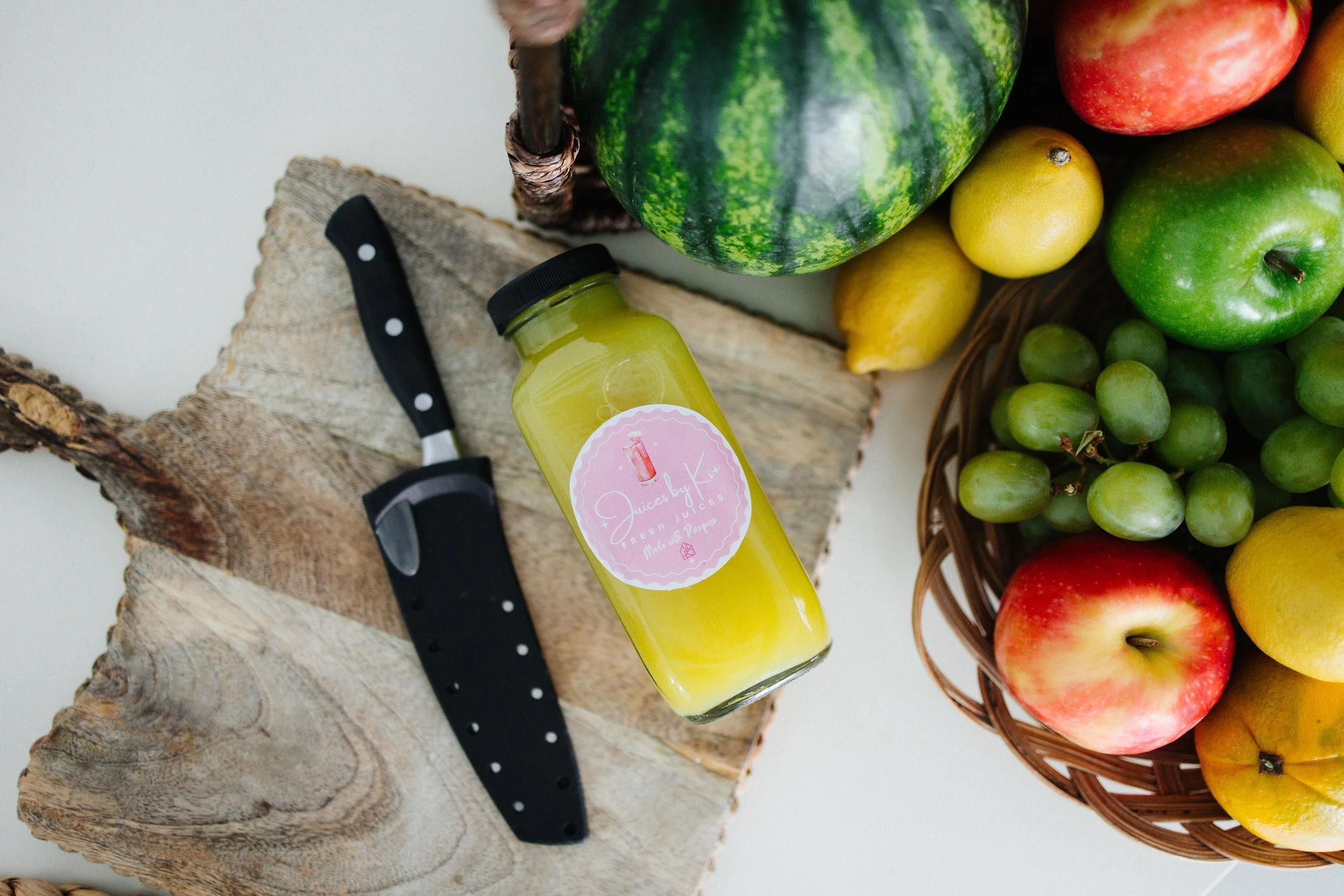 A bottle of yellow juice labeled "Juice by K", a black knife with white polka dots, a watermelon, some lemons, apples, and a bunch of green grapes in a basket, all on a wooden surface.