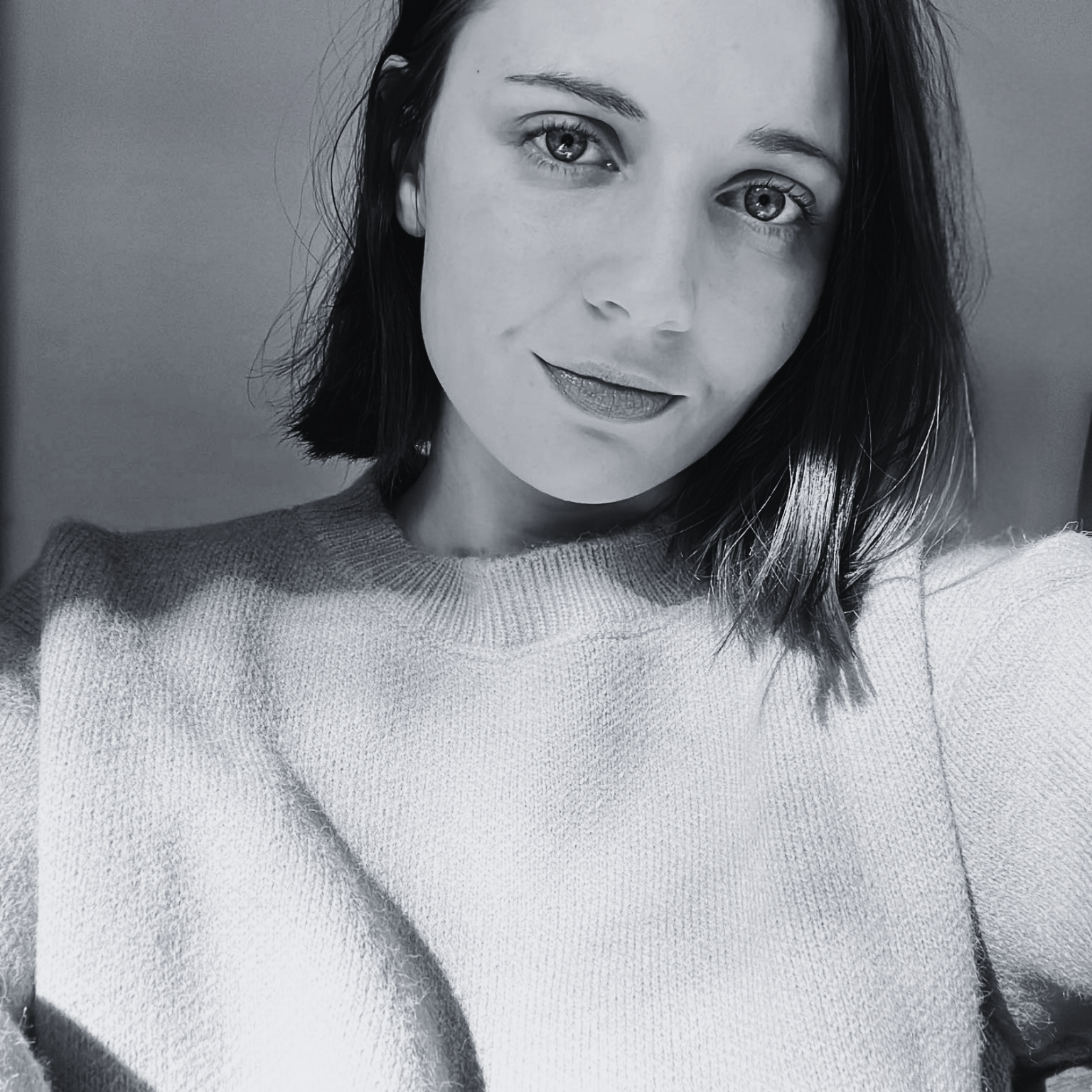 A black and white close-up selfie of a woman with shoulder-length dark hair, smiling slightly, wearing a light-colored sweater.