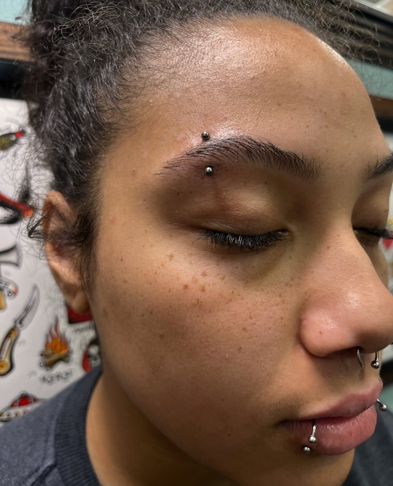 Close-up of a woman's face with closed eyes, multiple facial piercings, and visible freckles.