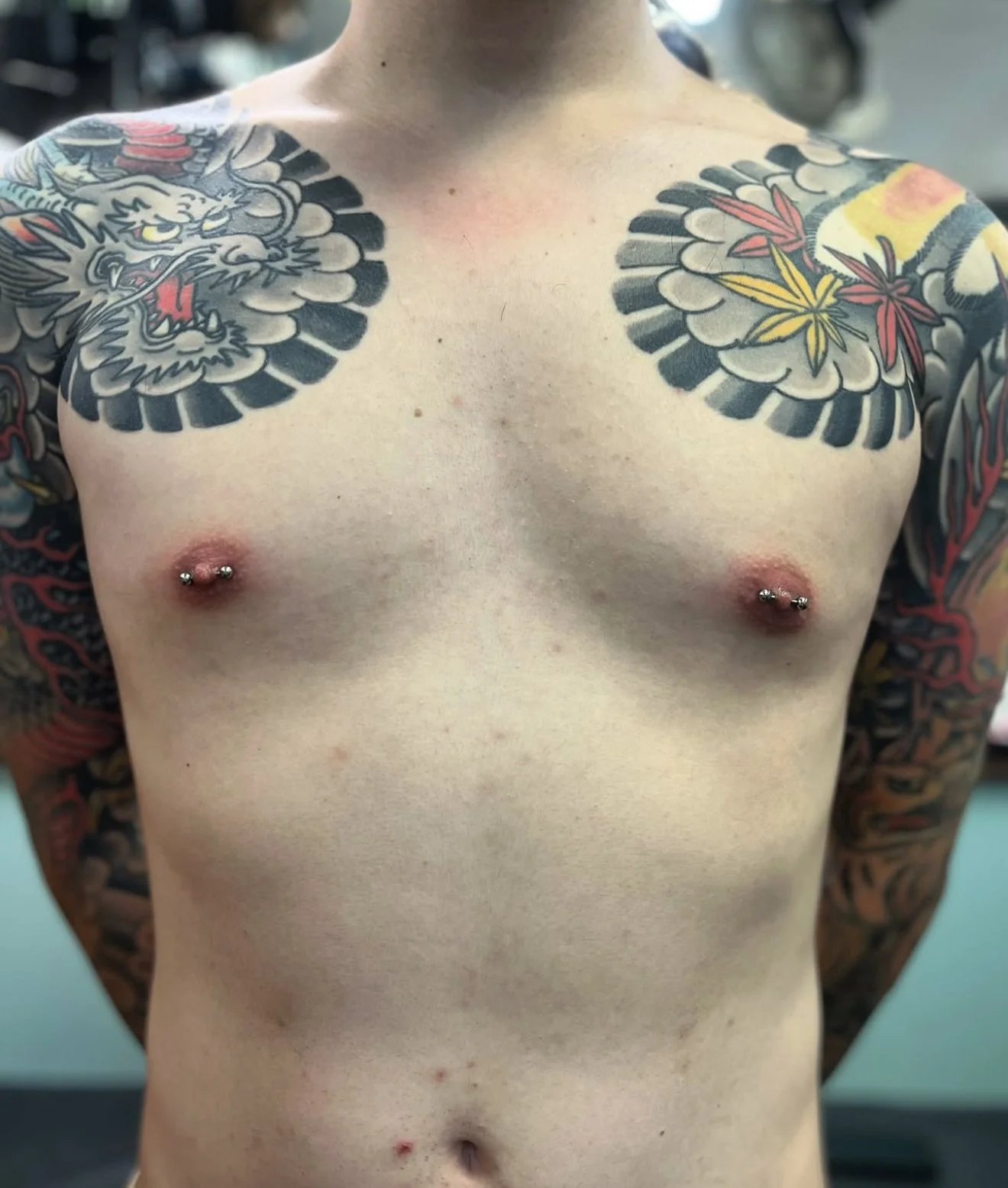 A shirtless person with colorful tattoos on the shoulders and arms, and nipple piercings.