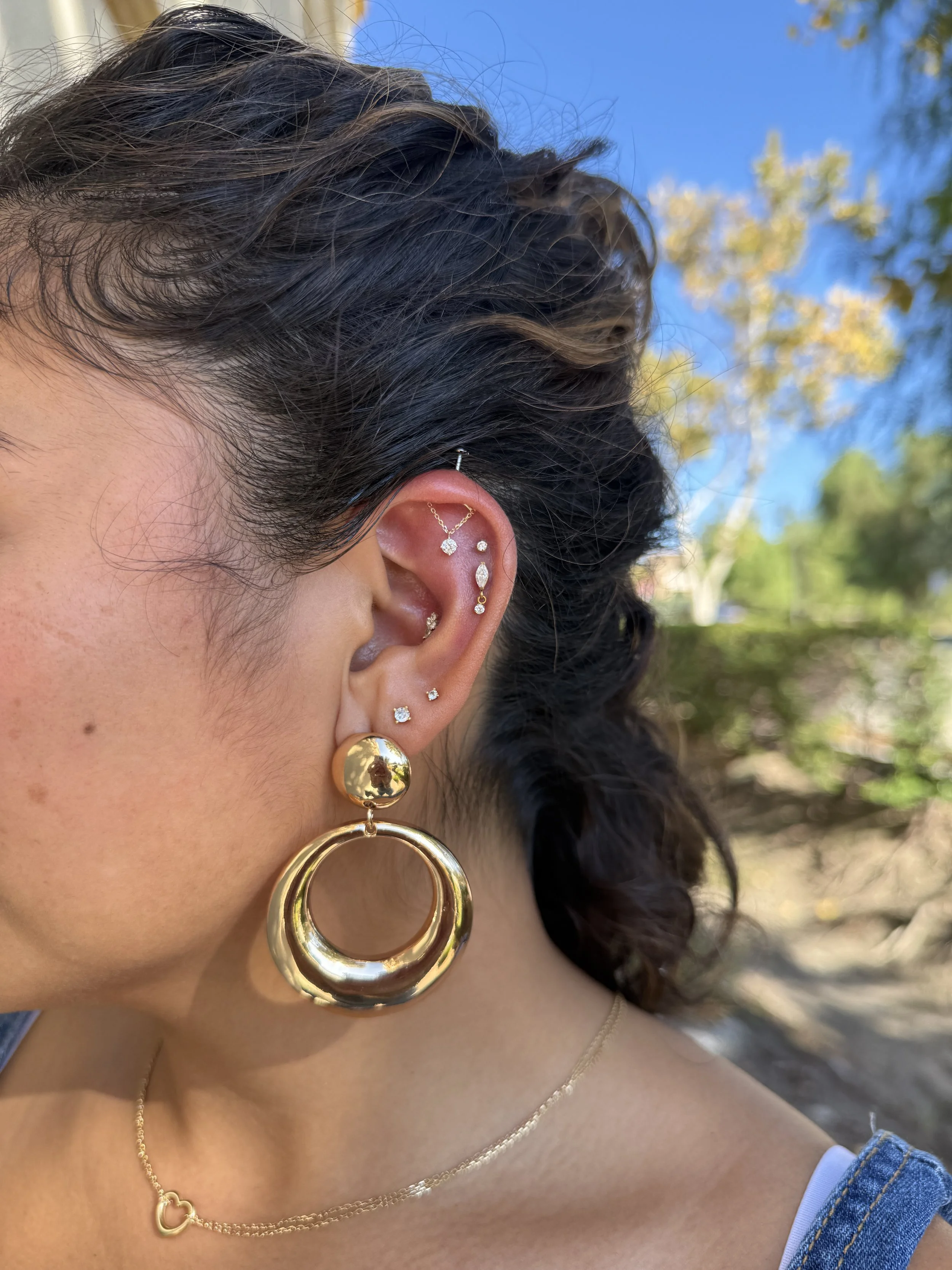A woman with dark curly hair showing multiple earrings in her ear and wearing a gold necklace with a heart shape pendant, outdoors with blue sky and trees in the background.
