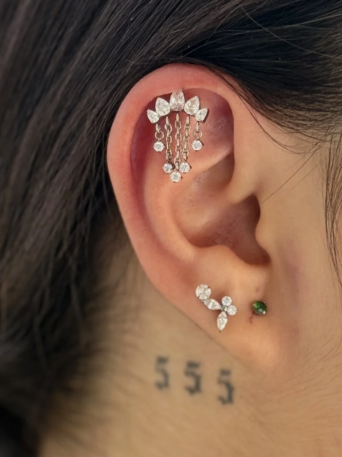 Close-up of a person's ear with decorative jewelry including a large ear cuff with clear stones and dangling chains on the upper ear, a star-shaped earring with multiple white stones on the lower earlobe, and a small green stud earring next to it. Part of a tattoo with the number 555 is visible on the neck.