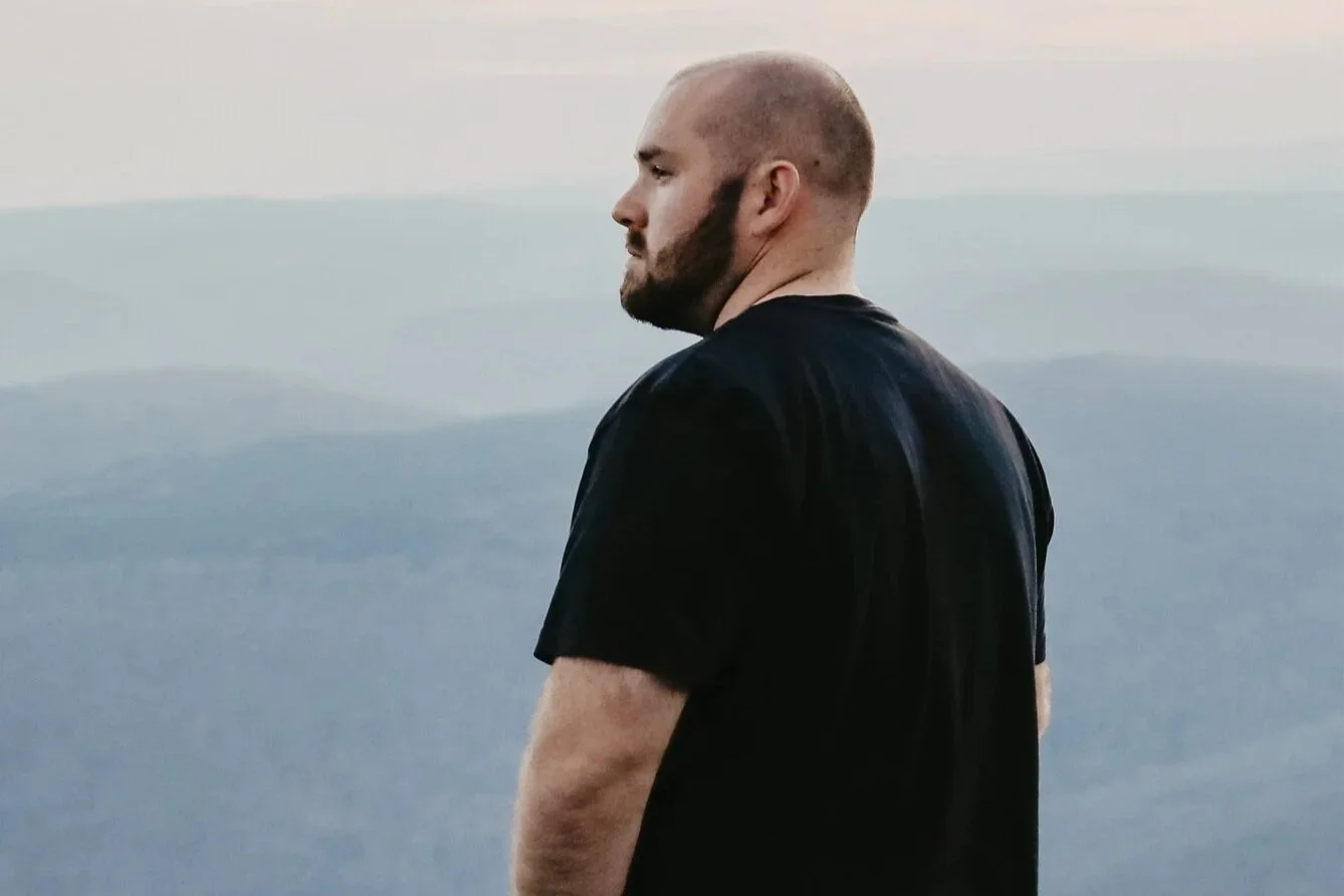 A man with a beard and shaved head is standing outdoors on a hill or mountain, wearing a black t-shirt and blue jeans, looking to the left with mountains and a cloudy sky in the background.