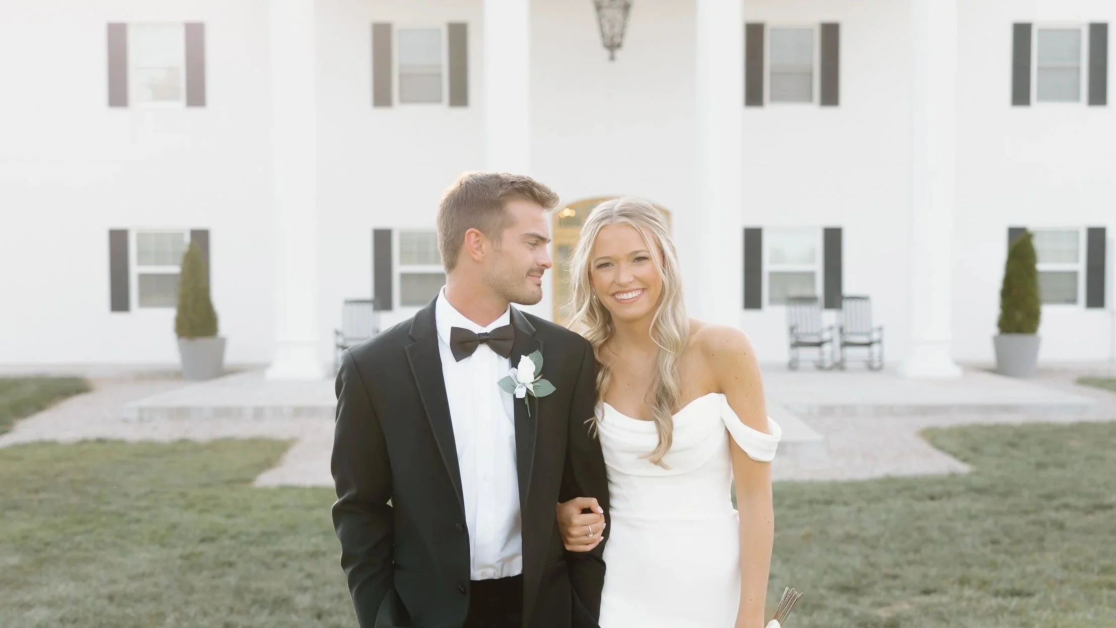 A bride and groom walking arm-in-arm on a grassy lawn in front of a white, colonial-style building with columns, black shutters, and potted plants, smiling and dressed in wedding attire.
