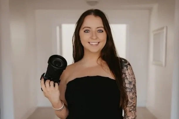 A woman with long brown hair and blue eyes smiling while holding a white flower on a stick, standing in front of a mirror with flowers in the background.