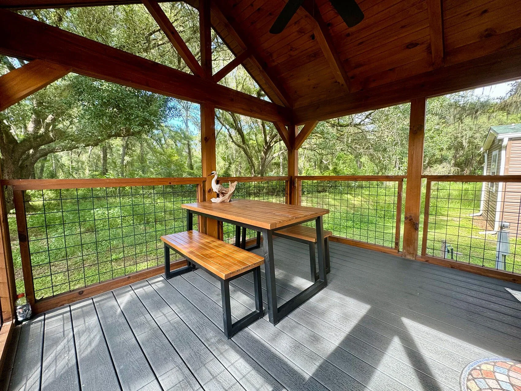 A cozy outdoor wooden porch with a rectangular table, a matching bench, and a decorative duck sculpture, surrounded by green trees.