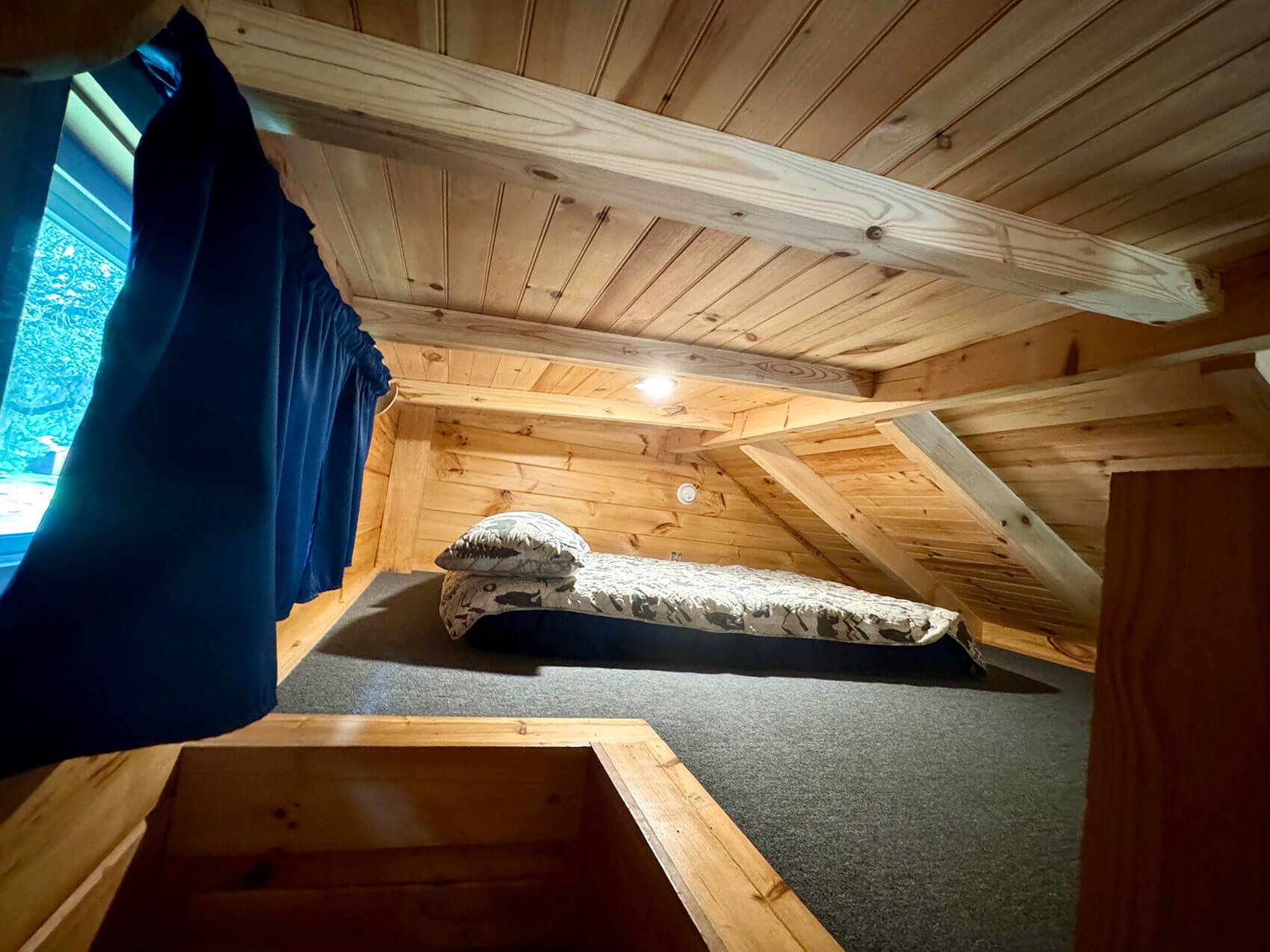 A cozy attic bedroom with wooden panel walls and ceiling, a single bed with a patterned comforter and pillow, a small window with blue curtains, and soft lighting.