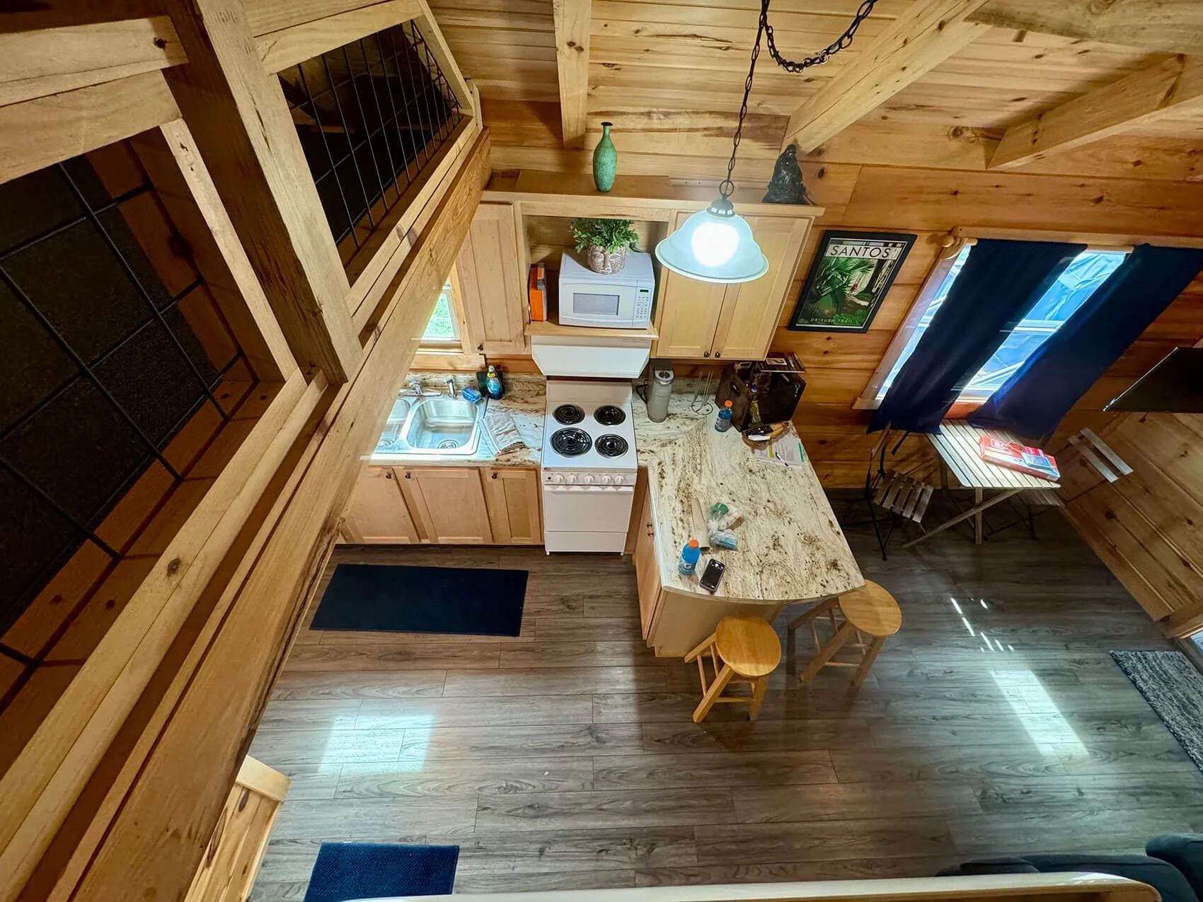 From an overhead view, a cozy kitchen and dining area with wooden walls and flooring, featuring a kitchen counter with a stove, microwave, kitchen sink, and stools at an island. Blue curtains hang over a window with a small table and two chairs nearb