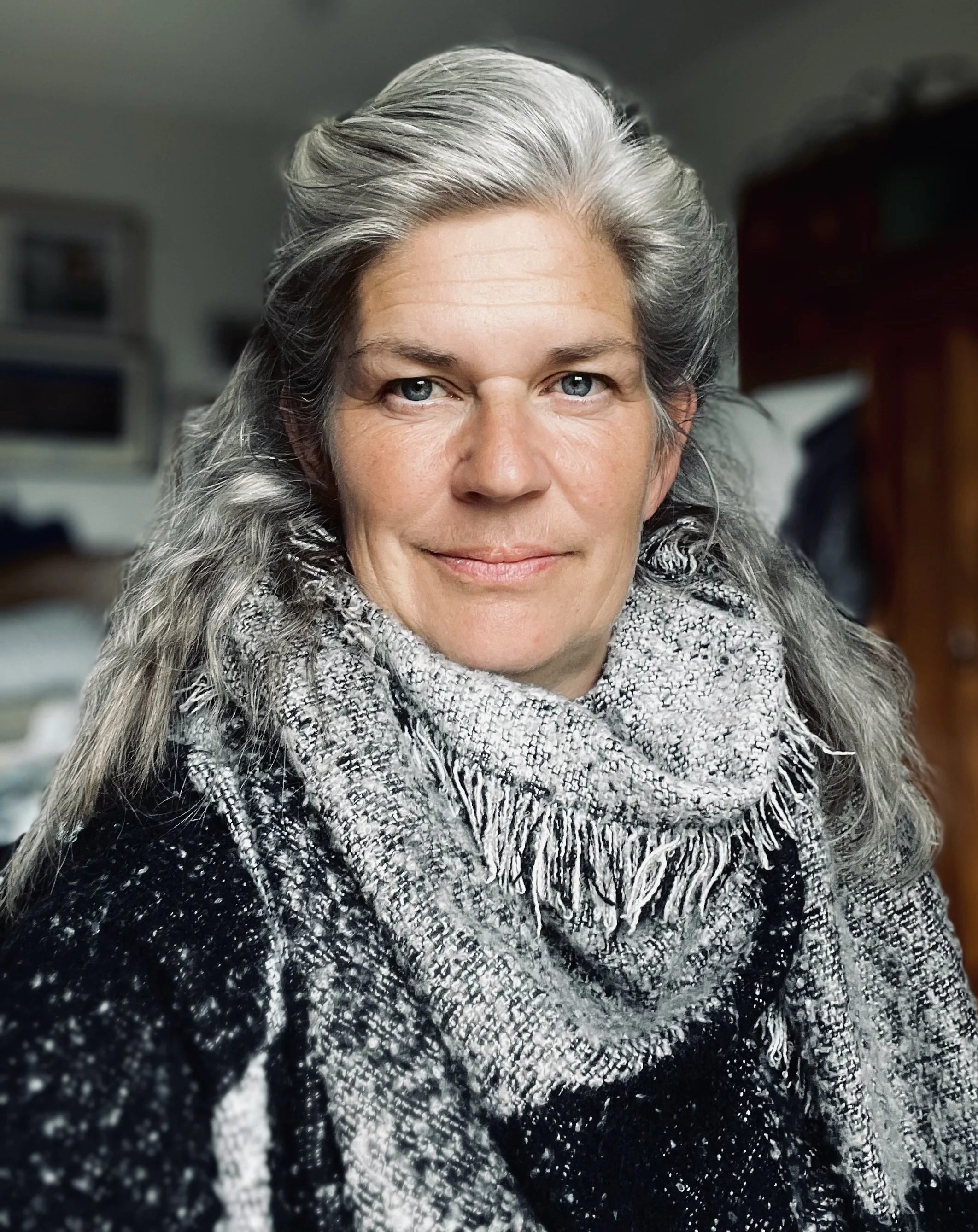 Close-up of a woman with gray hair and blue eyes wearing a black and white patterned scarf, smiling softly at the camera.