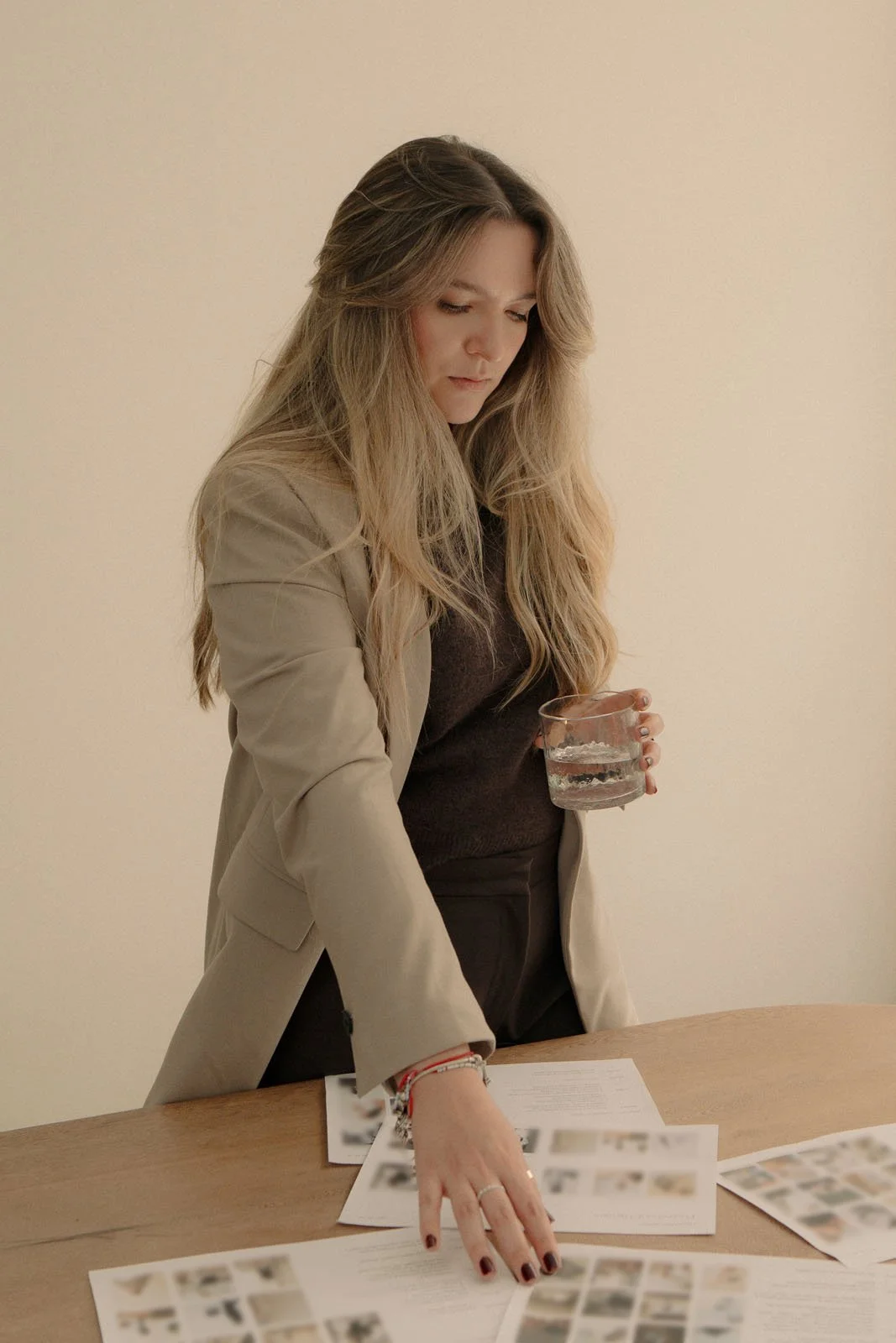A woman with long wavy blonde hair, wearing a beige blazer and dark top, is standing at a table with printed photos and documents, holding a glass of water.