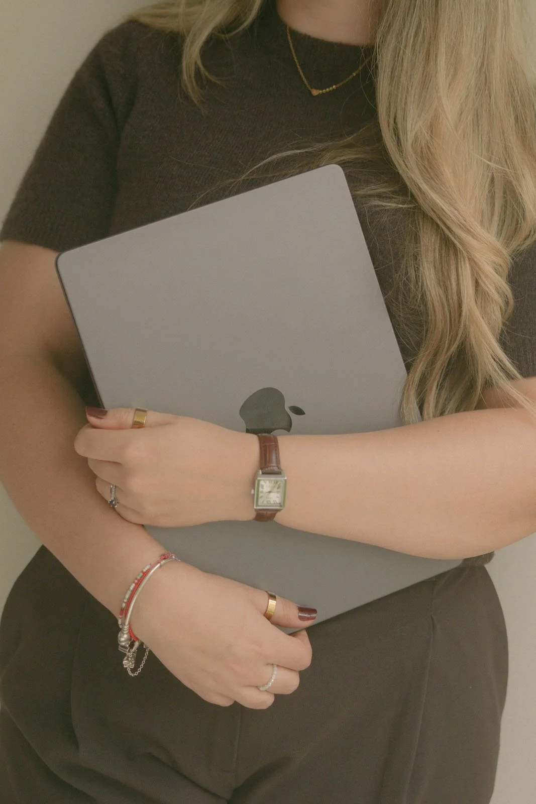 A woman with long blonde hair, wearing a black short-sleeved top, holding a closed silver MacBook laptop with an Apple logo, wearing a watch, rings, and bracelets on her left wrist.