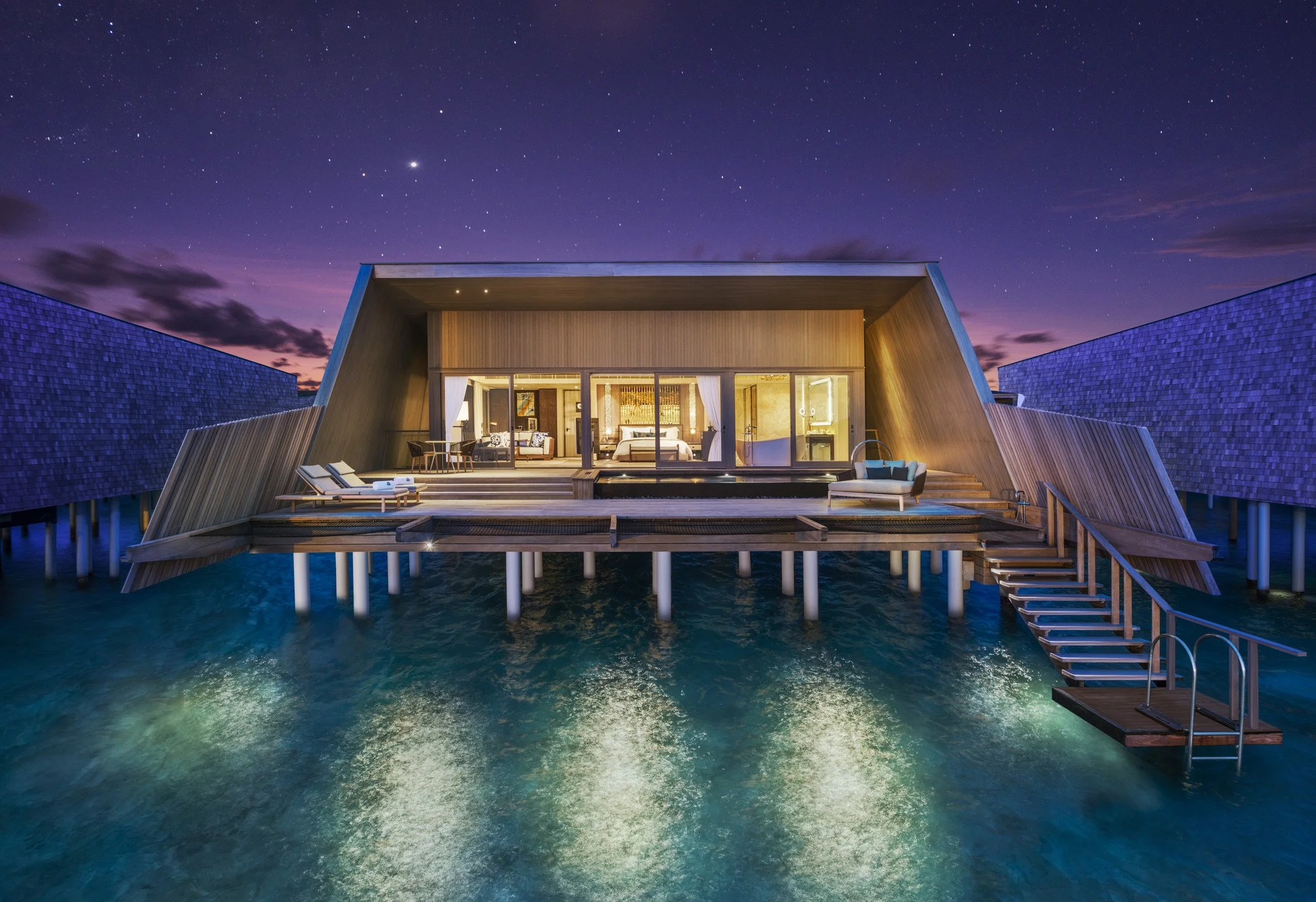Overwater luxury resort villa at night with illuminated interior, wooden deck, lounge chairs, and staircase leading into water, under a starry sky.
