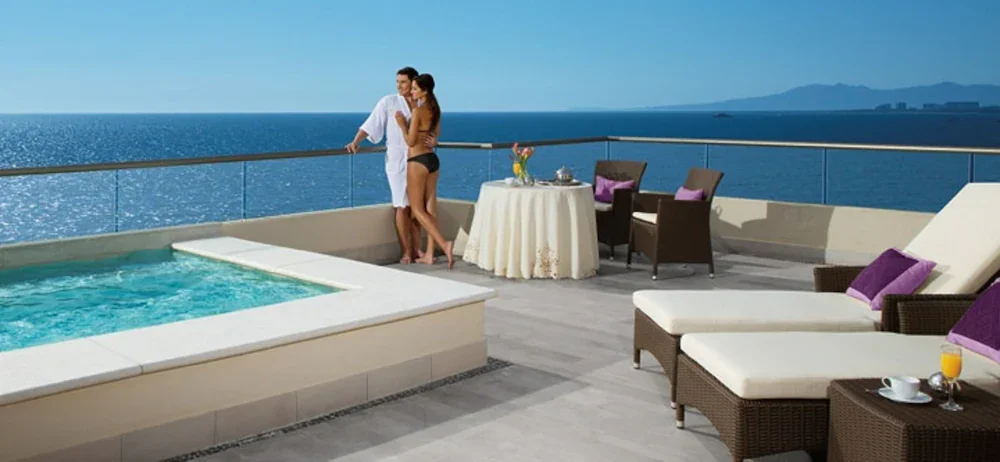 Couple standing on a rooftop terrace overlooking the ocean with a hot tub and outdoor seating, in a sunny setting.