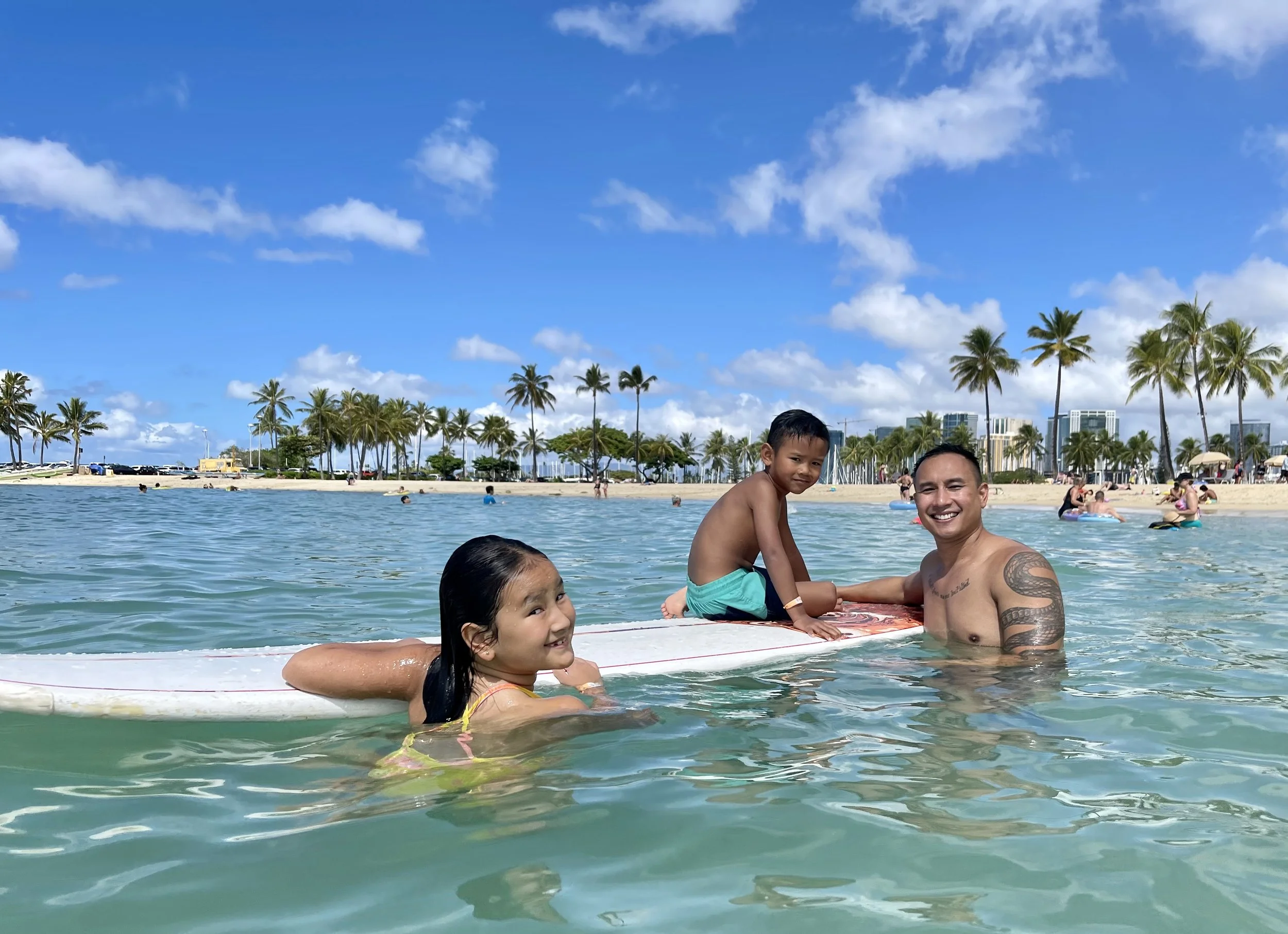 A man and two children enjoying time in the water at a beach with palm trees, blue sky, and people in the background.