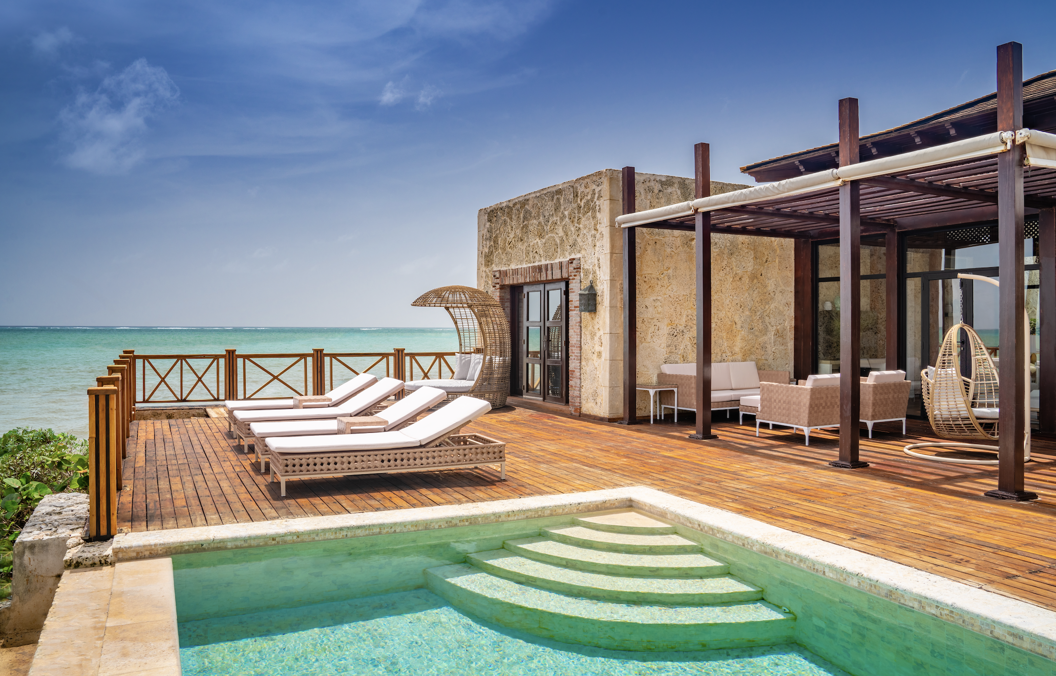 Beachside pool area with lounge chairs, a shaded seating area, and a view of the ocean under a blue sky.