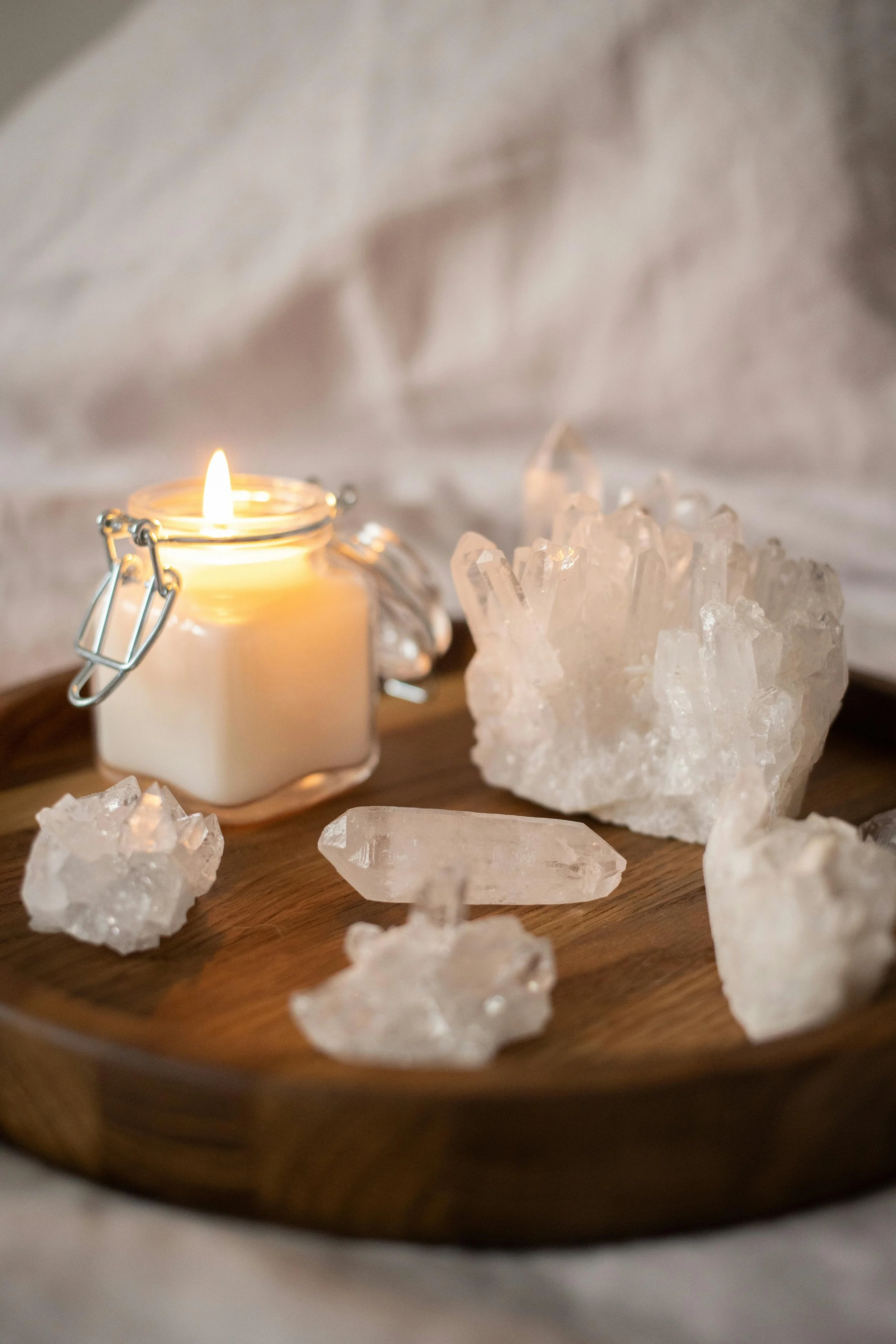 A lit candle in a jar next to various clear quartz crystals on a wooden tray.