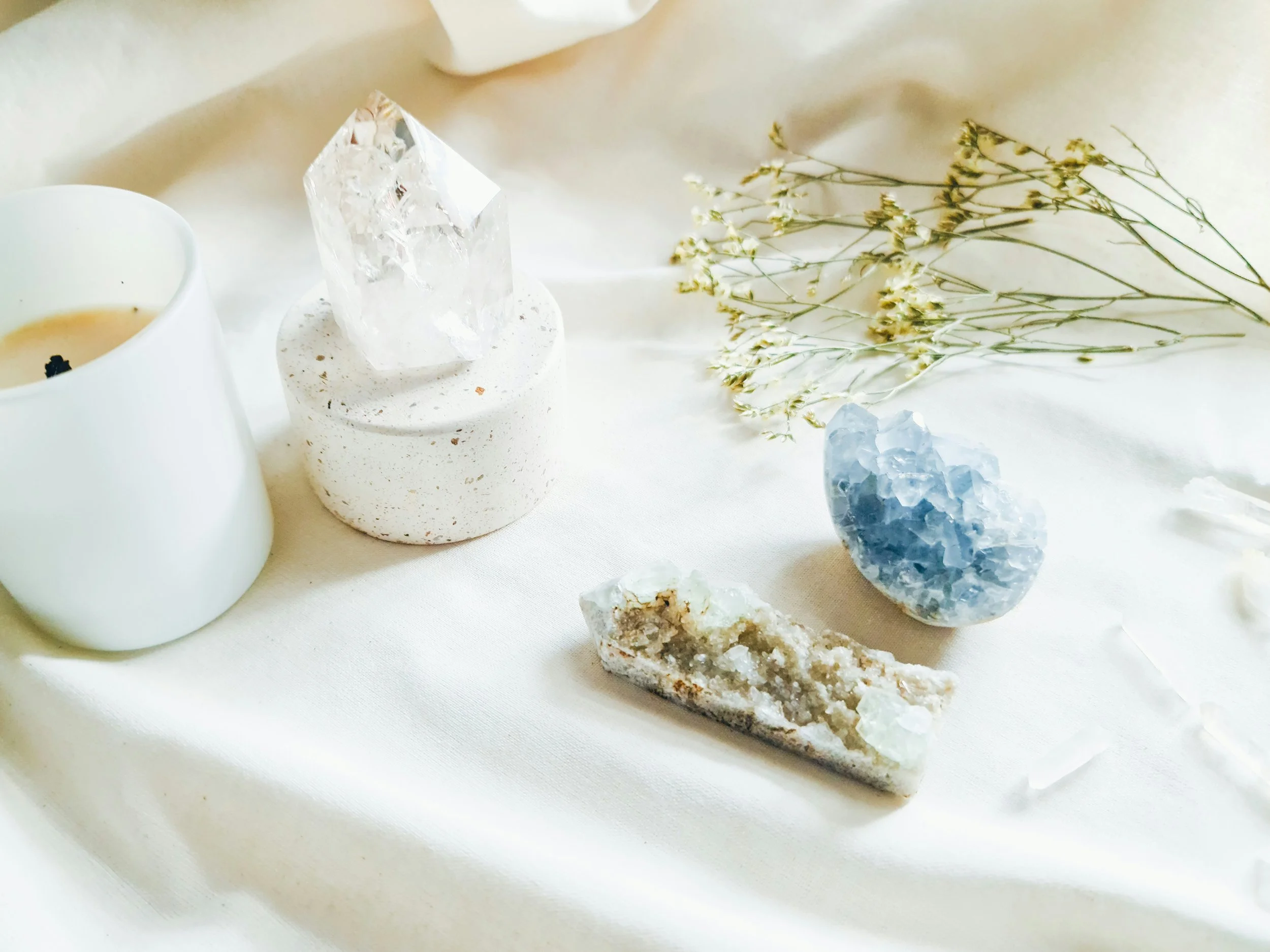 Decorative arrangement of crystals, dried flowers, and a candle on white fabric.