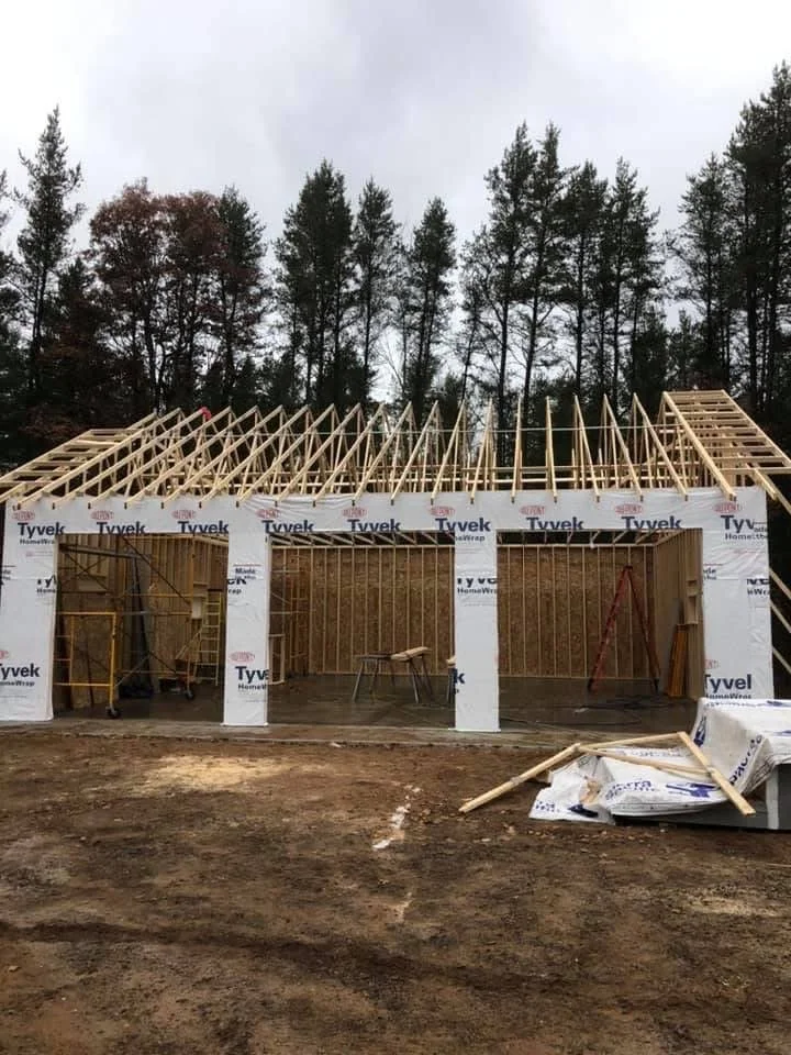 A house under construction with a wooden frame and roof structure, surrounded by trees and a cloudy sky.