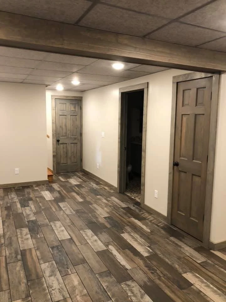 Empty room with wood-look tile flooring, beige walls, and three wooden doors, one open to a bathroom, with ceiling lights.