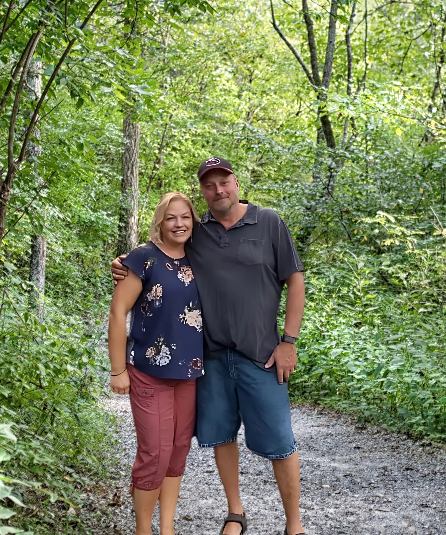 A man and woman smiling outdoors on a forest trail, arms around each other, surrounded by green trees.