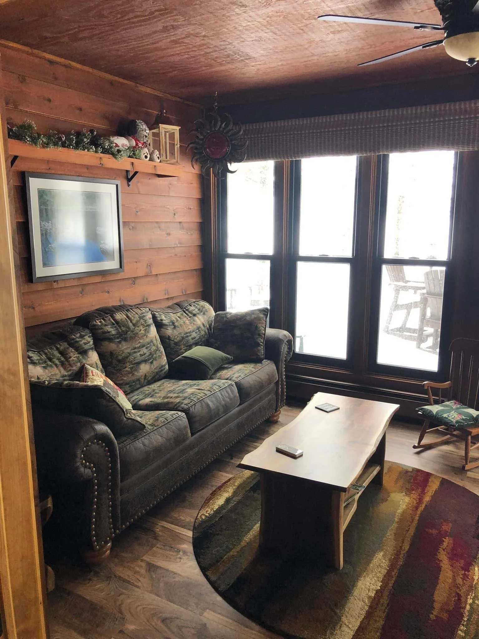 Cozy living room with wooden walls, a patterned sofa, a wooden coffee table, a window with natural light, and holiday decorations on a shelf.