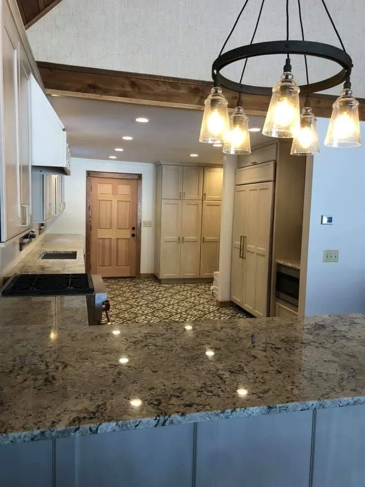 Modern kitchen with granite island counter, cream-colored cabinets, patterned floor tiles, built-in oven, and hanging light fixture with exposed bulbs.
