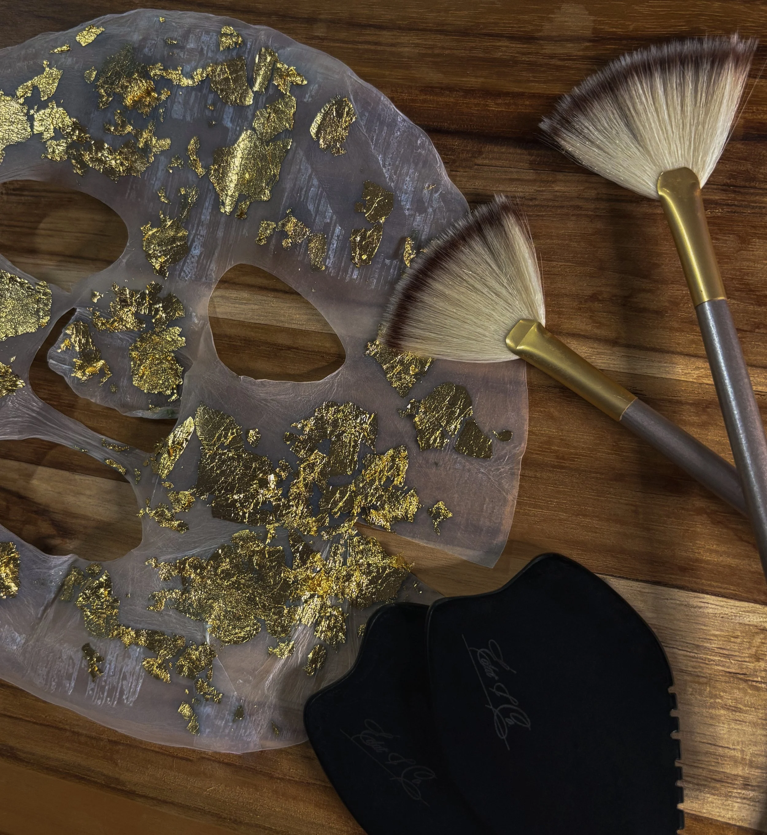 A translucent face mask decorated with gold leaf, placed on a wooden surface, with two makeup brushes next to it. One brush has a gold handle and the other has a dark handle with cursive writing.