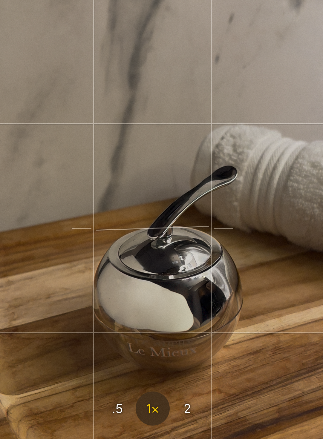 A shiny silver ball-shaped container with a small handle on top, labeled "Le Mieux", is placed on a wooden surface. A rolled white towel is behind it, against a light-colored wall.