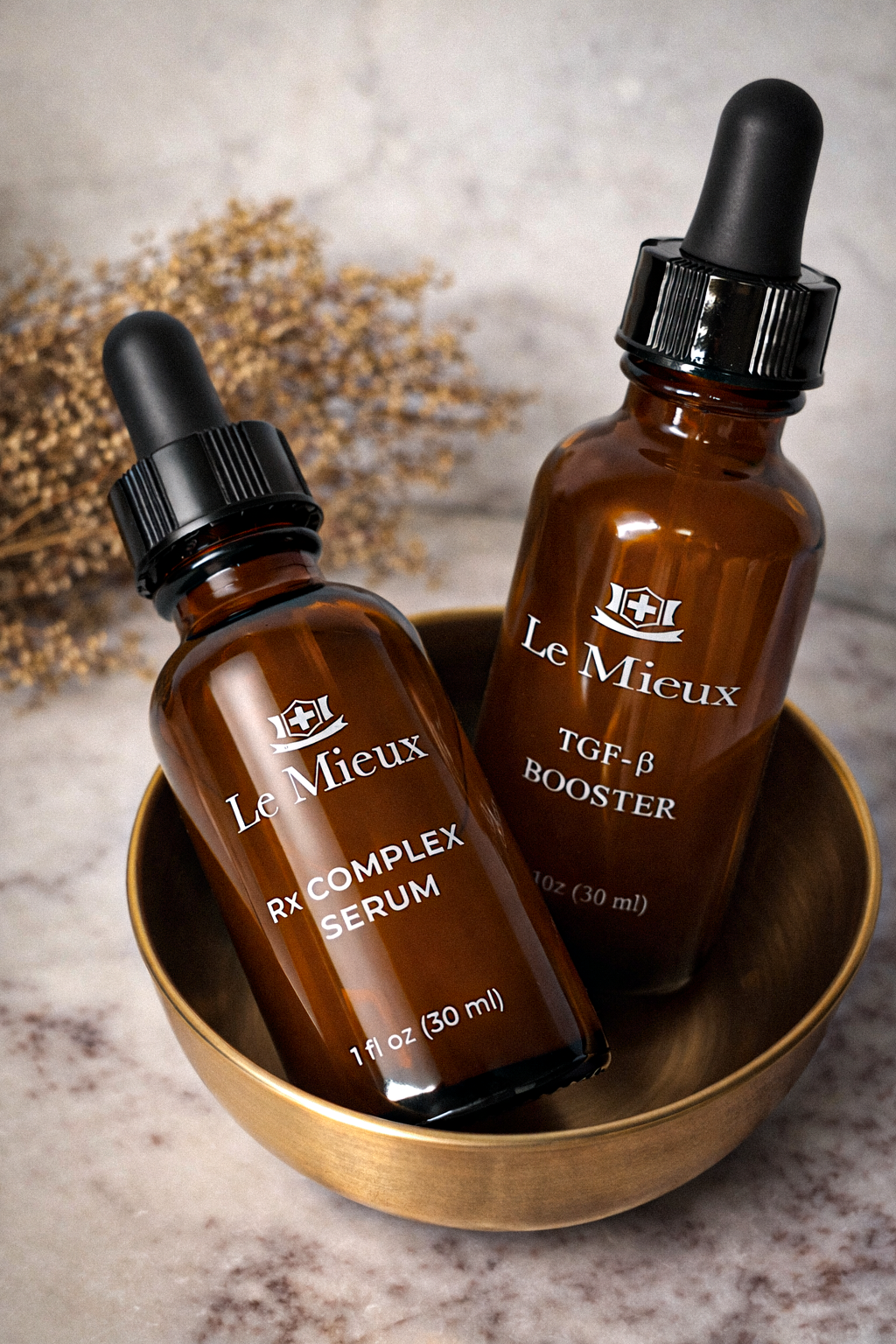 Two amber glass dropper bottles labeled Le Mieux RX Complex Serum and TGF-β Booster placed in a gold bowl, with dried plants in the background.