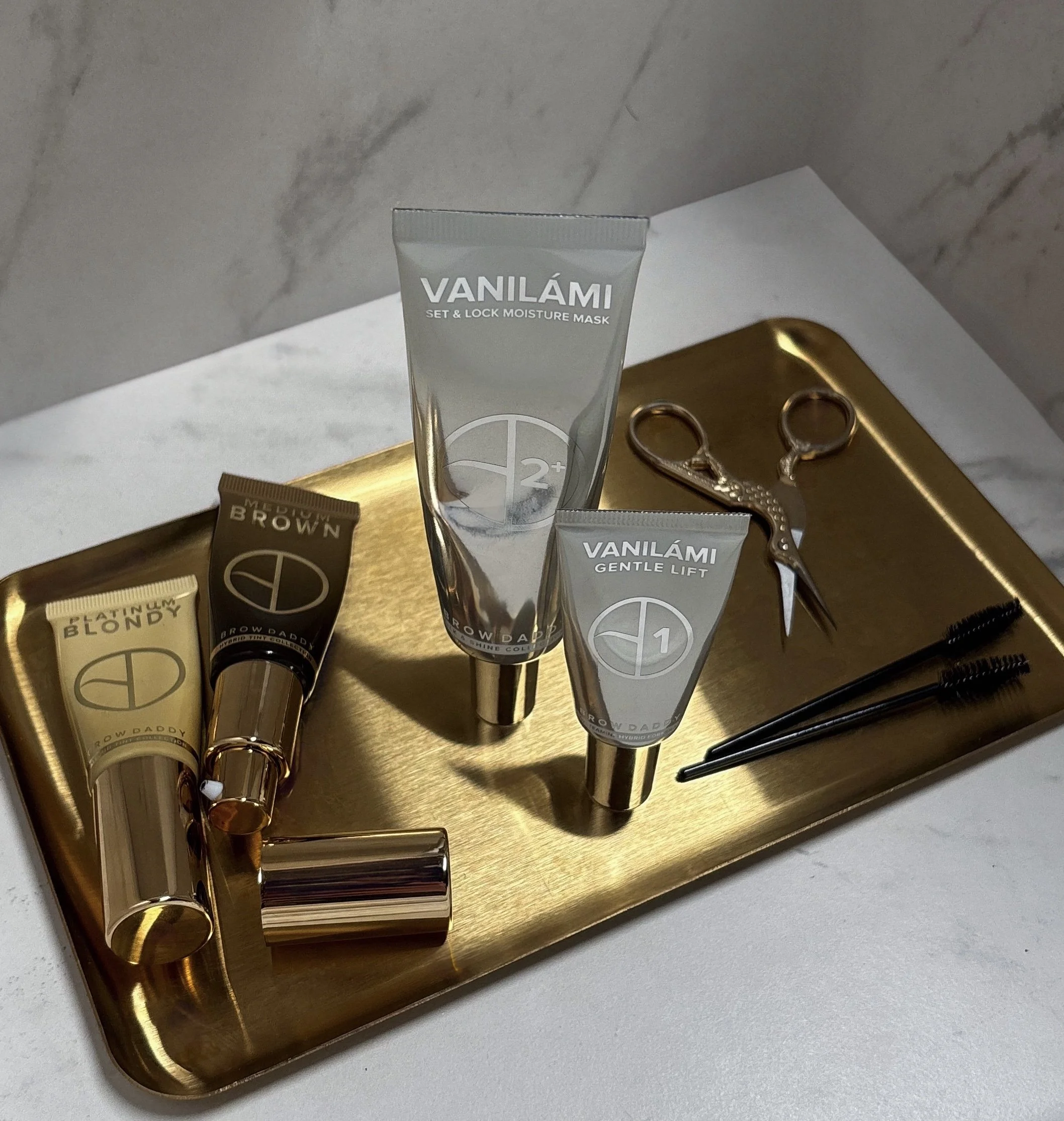 Beauty products including tubes labeled Vaníami and other skincare items arranged on a gold tray with scissors and a black hairbrush on a white surface.
