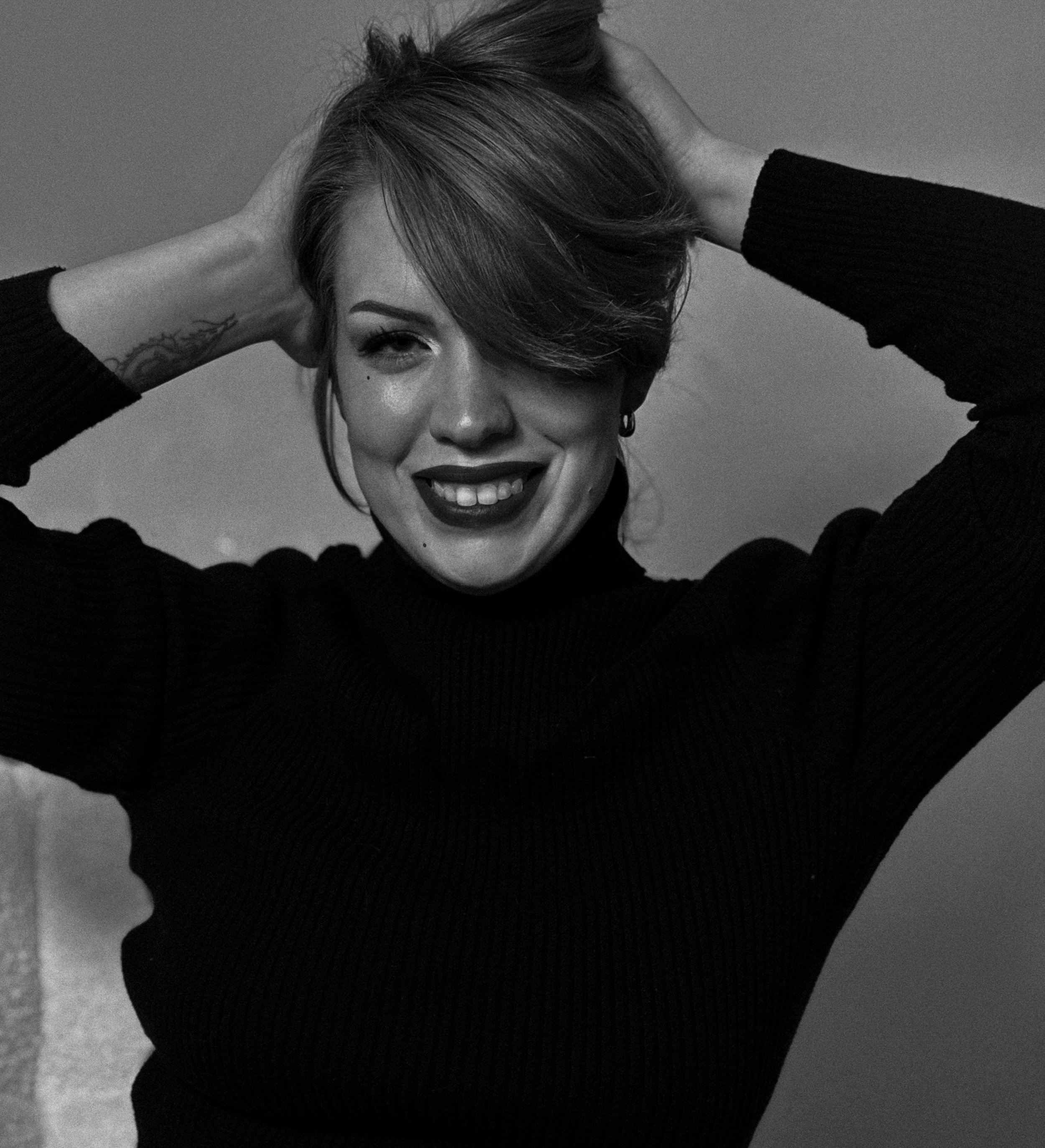 Black and white photo of a woman smiling with her hands behind her head, wearing a long-sleeve sweater, with short hair and a tattoo on her right wrist.