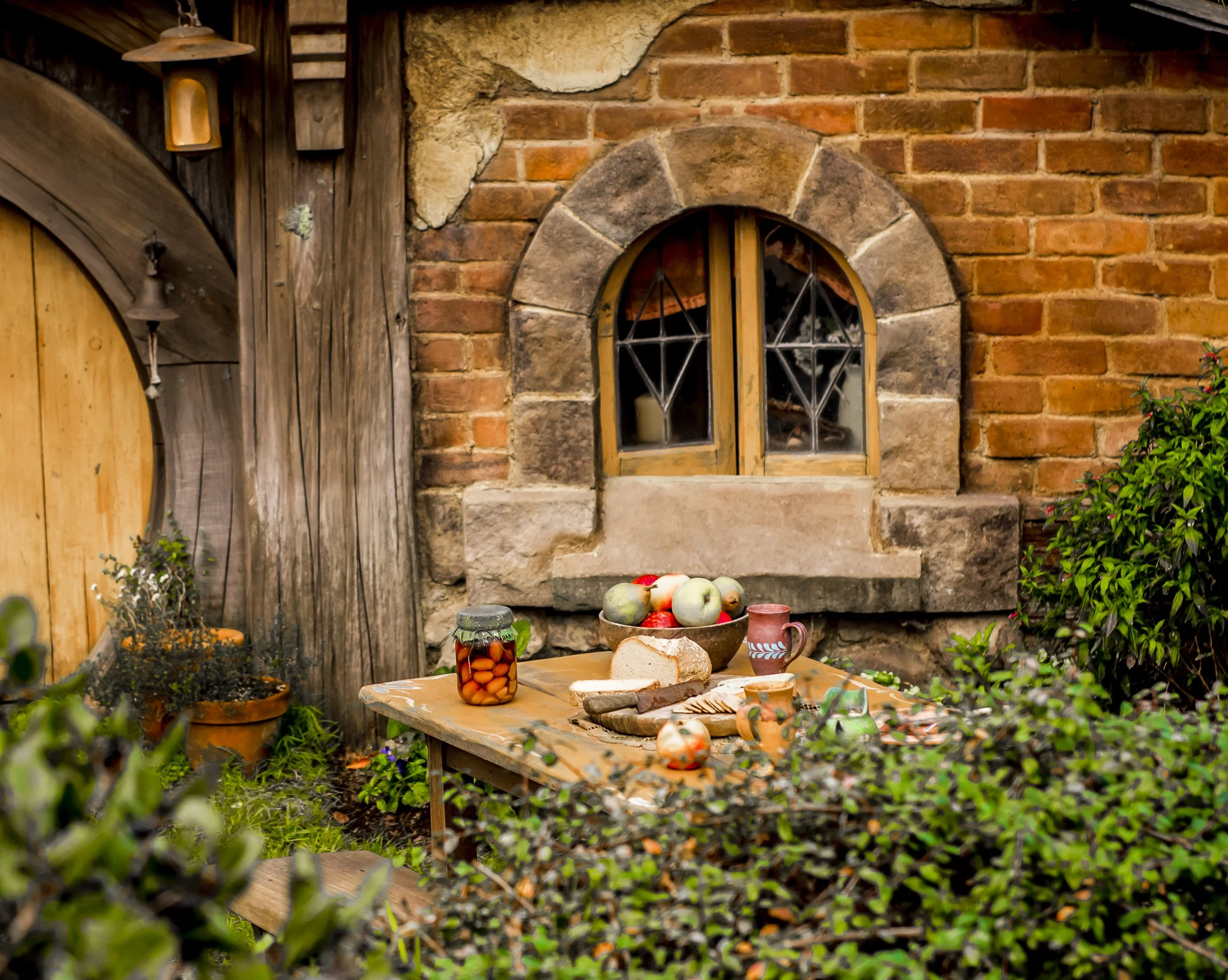 A cozy little Hobbit table in front of a Hobbit hole, all set with lots of yummy looking bread, cheese, and fruit.