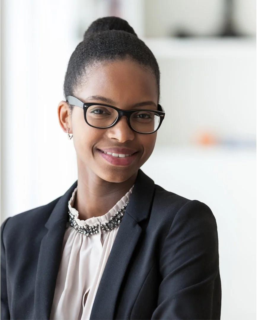 A smiling African American woman in business attire, wearing black glasses, a black blazer, and a pearl and black beaded necklace, standing in an office setting.